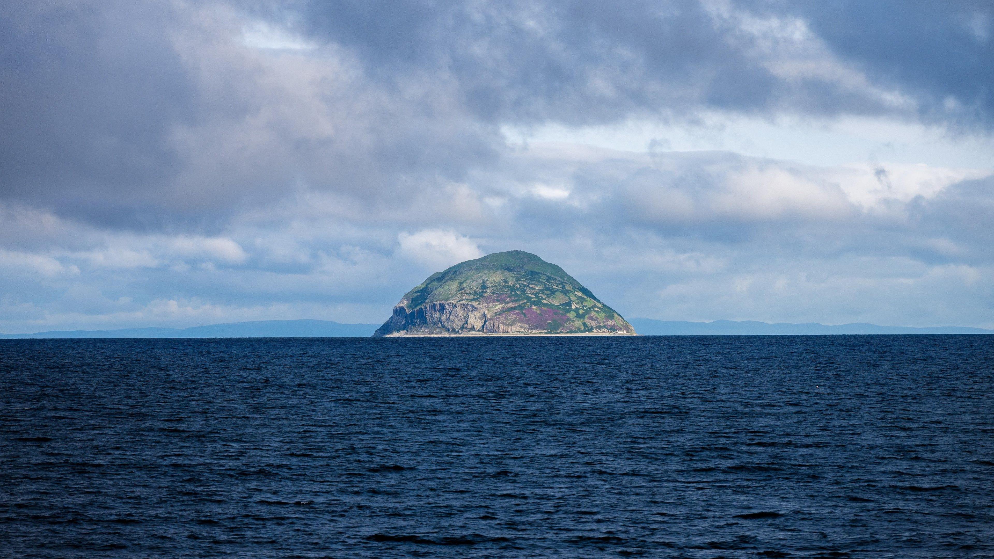 A view across dark water towards Ailsa Craig. The water is a dark blue colour. The island is brown and rocky with patches of green. The sky above is blue but streaked with white and grey clouds.