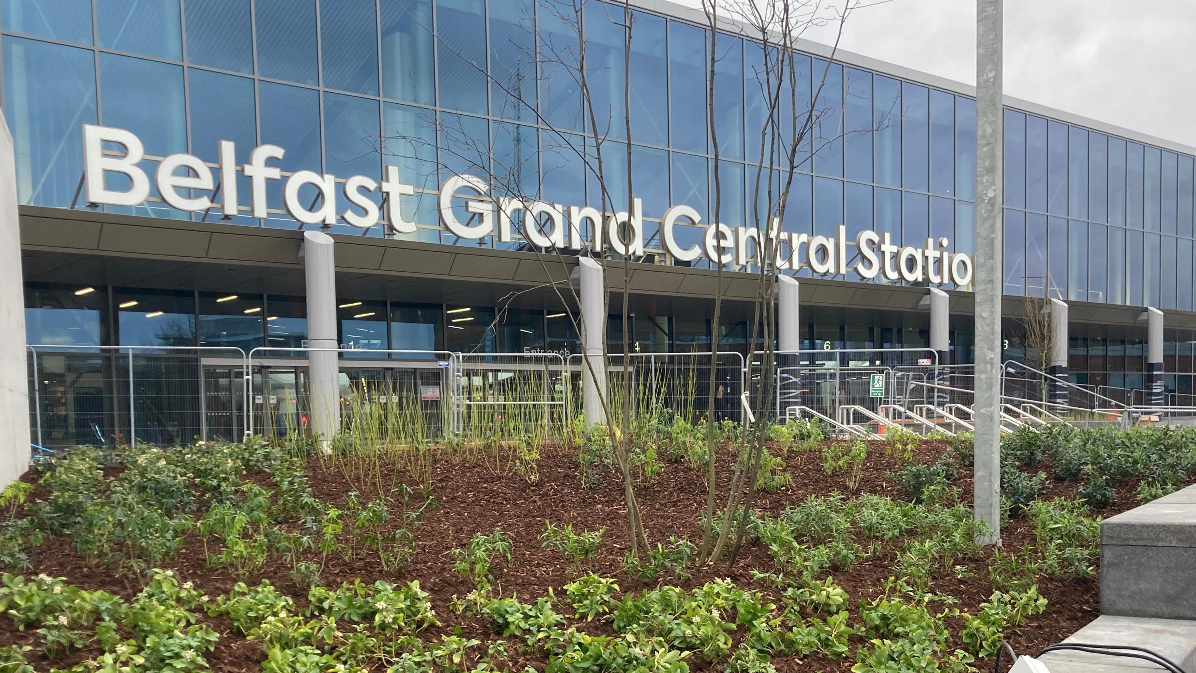 The station, with its transparent windows, sign in white letters which reads 'Belfast Grand Central Station' and green leafy plants in front. 