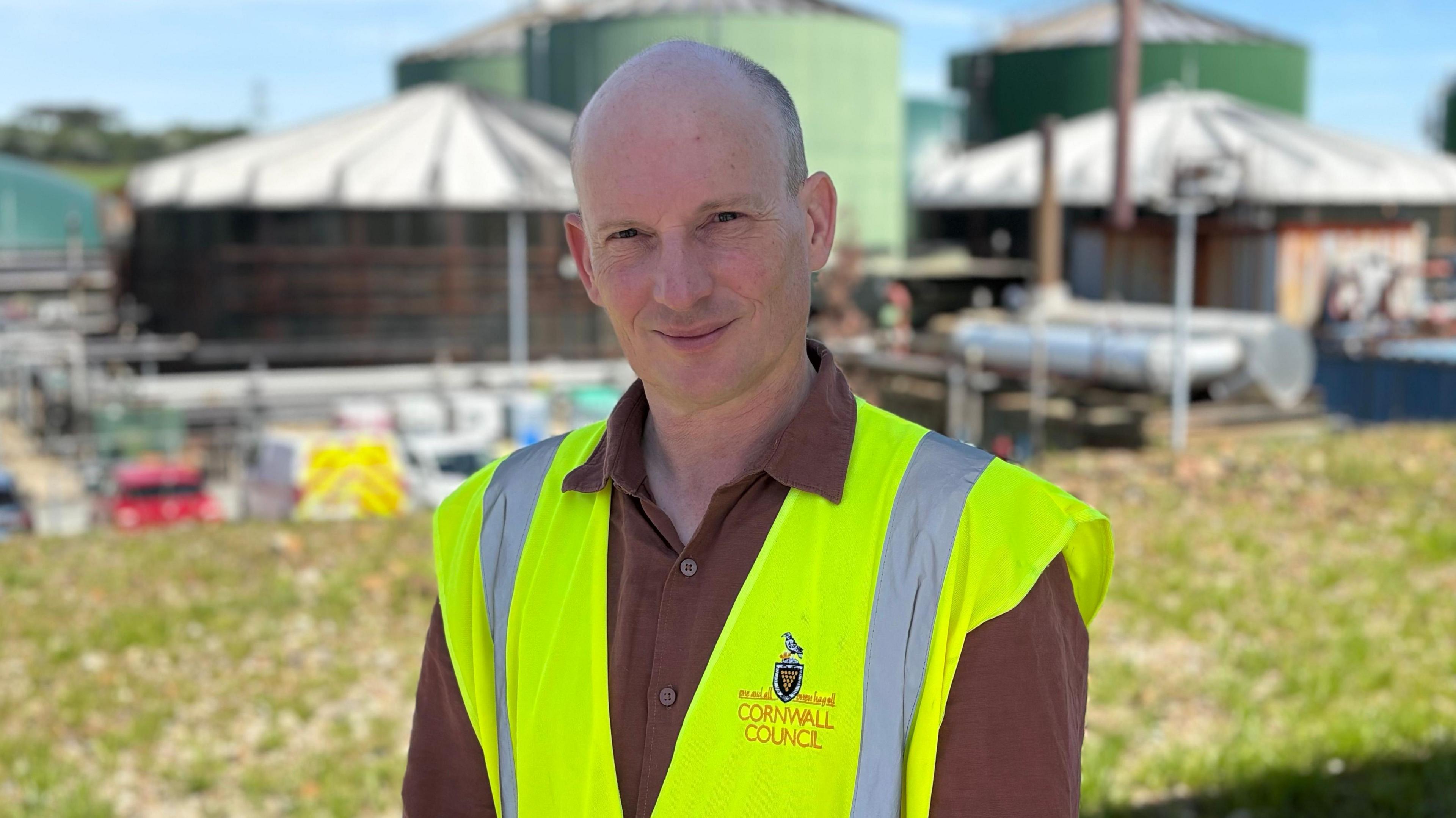 Loic has a shaved head and wears a red shirt underneath a yellow hi-vis vest with Cornwall Council's logo on it. He stands with the digester tanks behind him in the distance. 