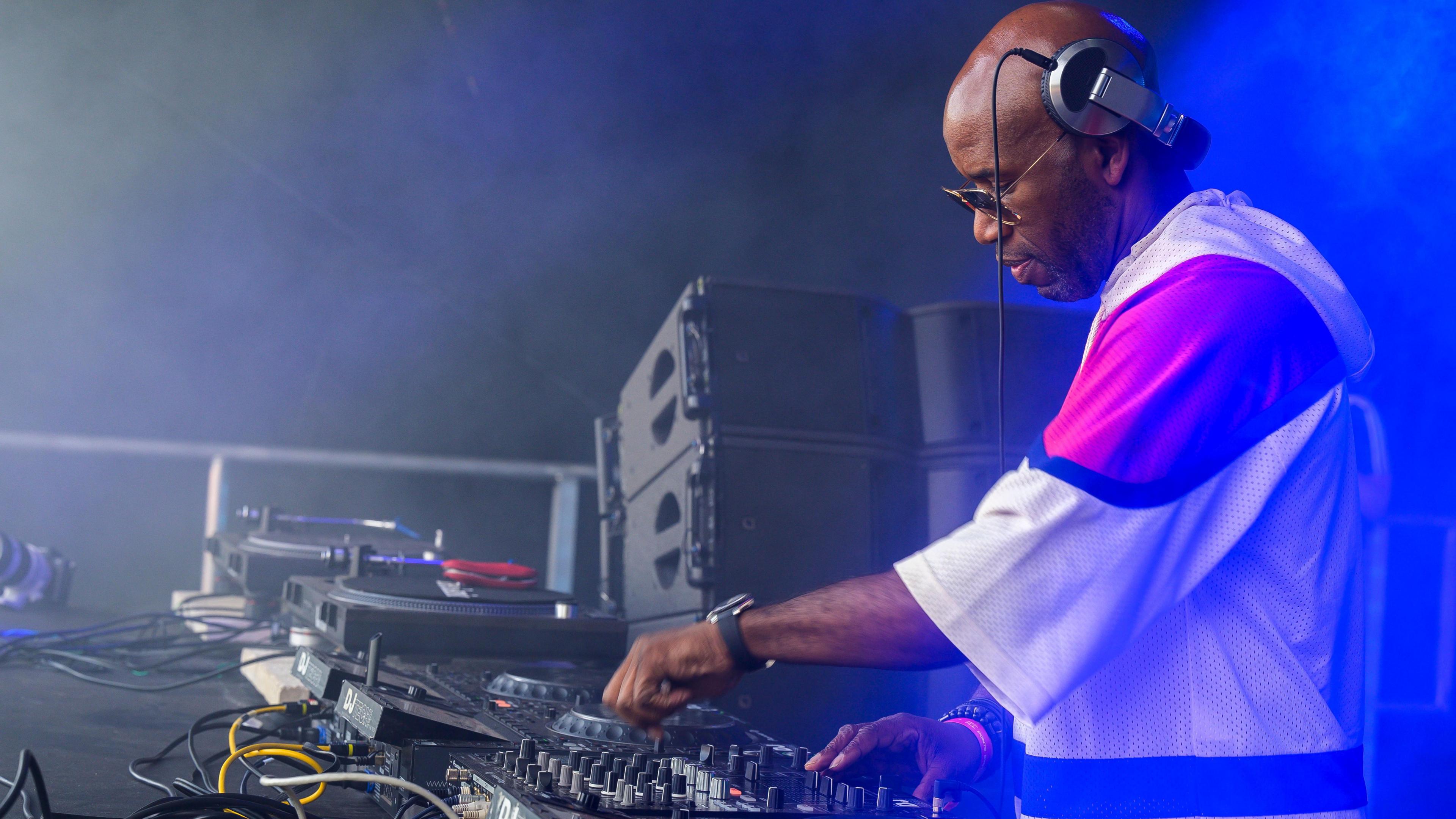 DJ Spoony, wearing baggy t-shirt, glasses and headphones is controlling a music mixing desk