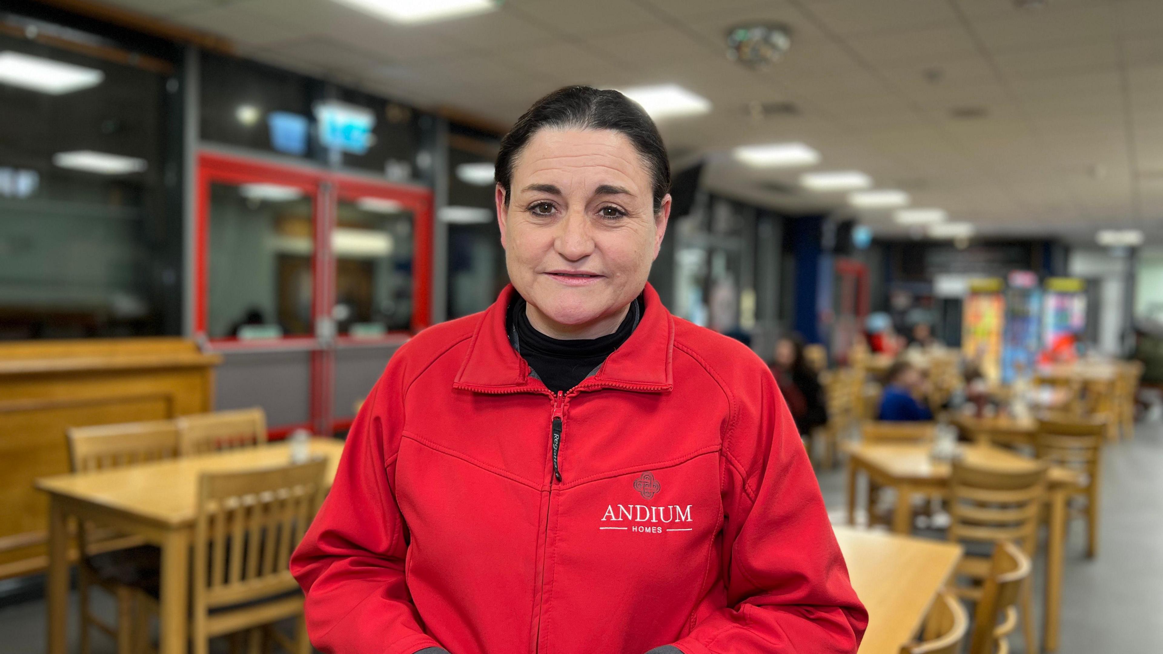 Dominique Caunce wears a red jacket branded with Andium Homes and faces the camera, with tables and chairs behind her.