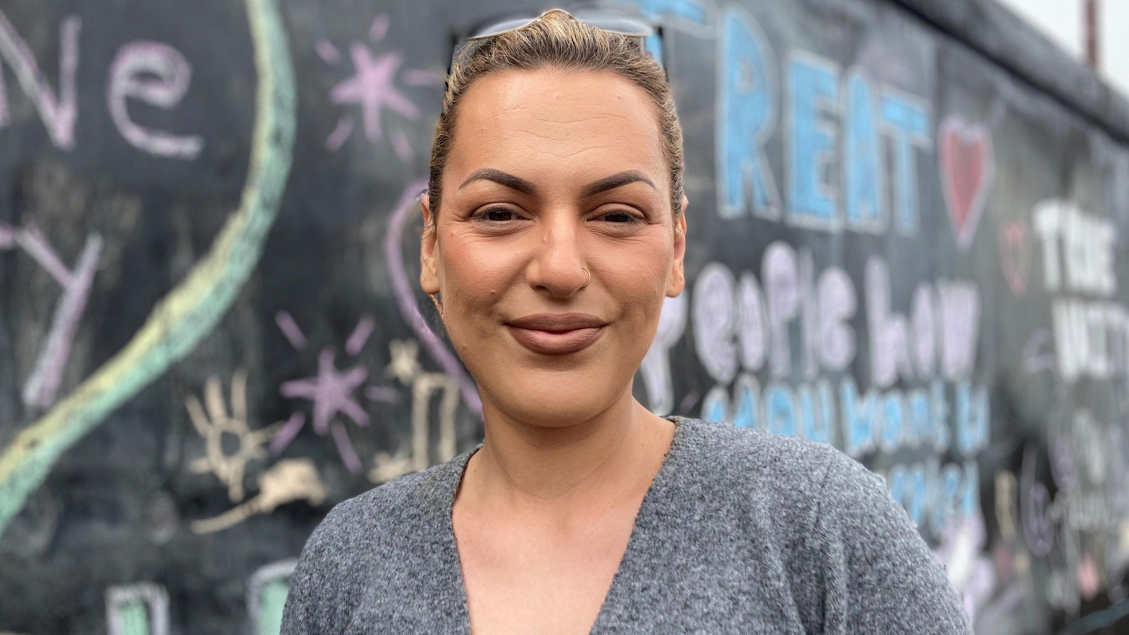 Anna Brown a woman who is standing in front of hoarding by a construction site. On the wall behind her is messages of support written in different colours of chalk. She is looking directly at the camera and is smiling. She is wearing a grey outfit and has blonde hair and a pair of glasses on the top of her head.