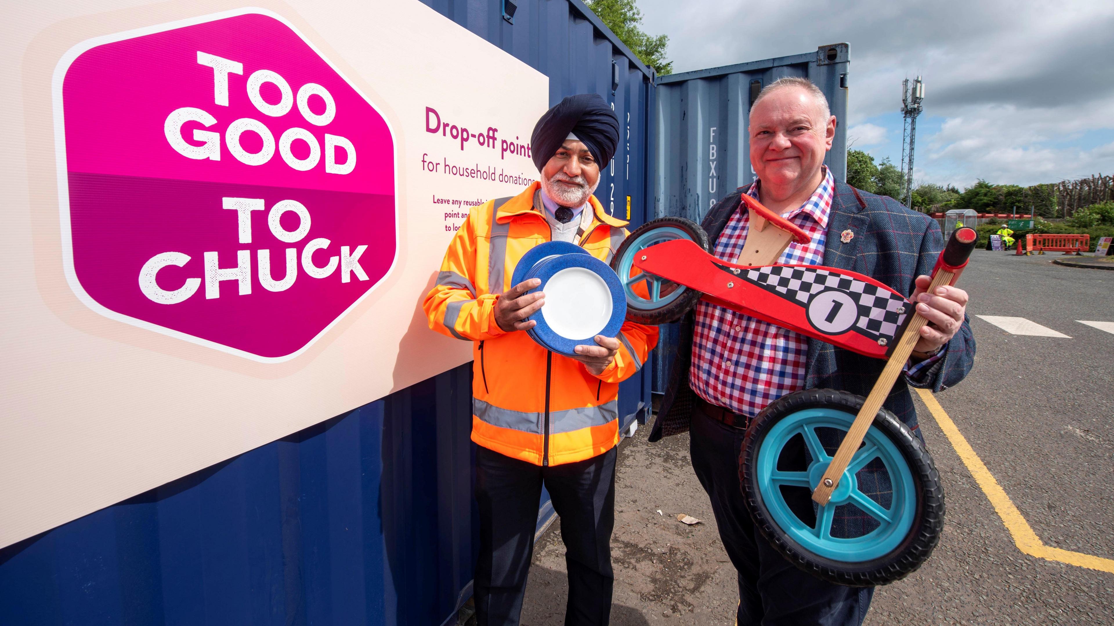 Two men, one holding plates and another holding a small, wooden kids bike, standing next to a blue shipping container at the side of a road. A pink posted is on the side of the container and says "Too Good To Chuck".