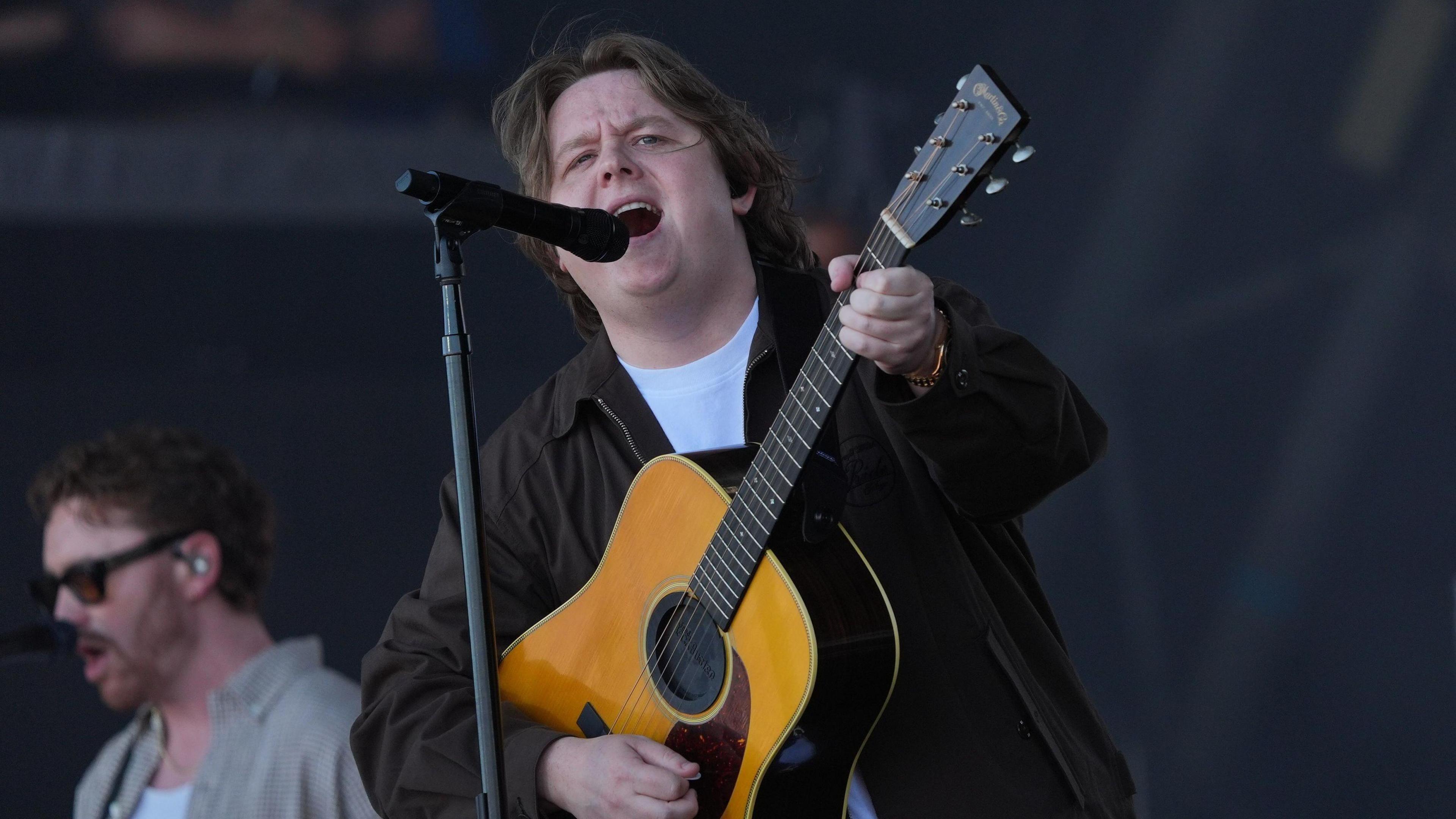 Lewis Capaldi is on stage with an accoustic guitar singing into a microphone. He has long hair and he is wearing a black jacket.