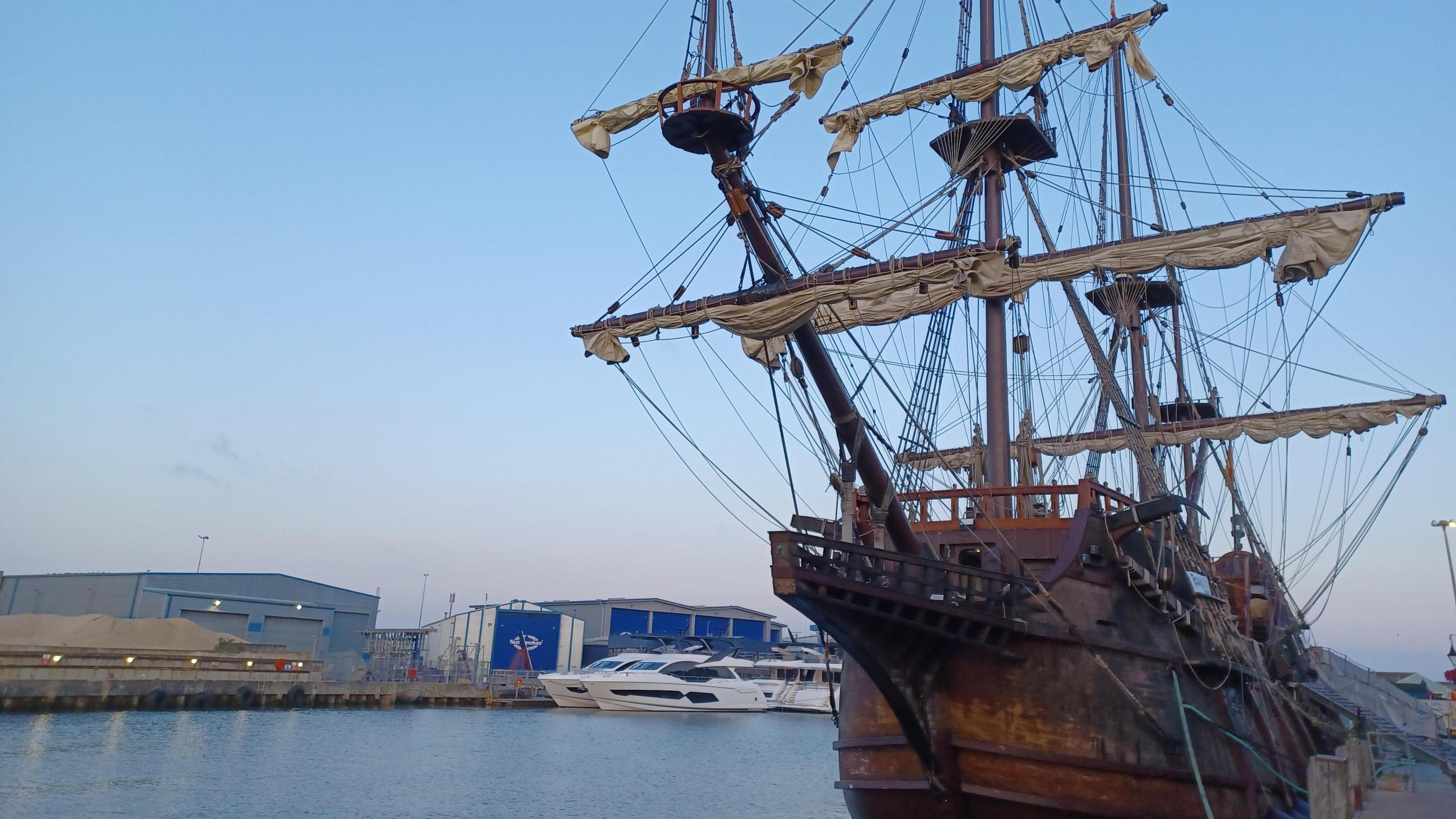 An old historical-looking sailing boat is in the foreground on a waterway with modern buildings in the background. The sky is clear and sunny