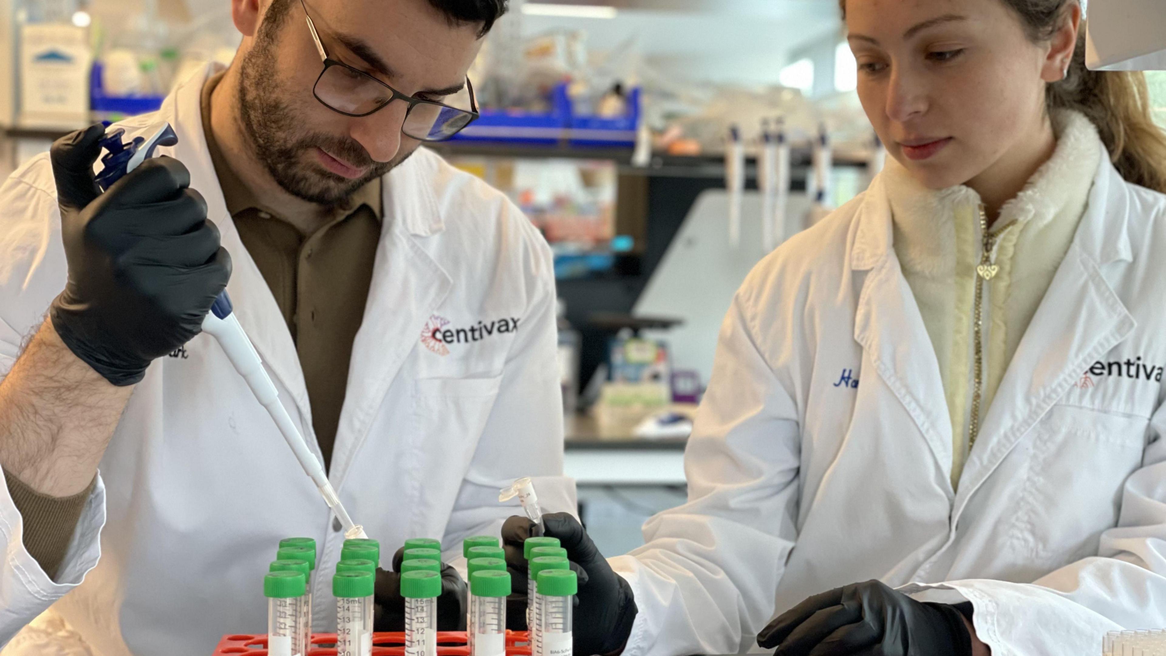 Two scientists wearing white lab coats and black gloves are pipetting into small vials in a laboratory