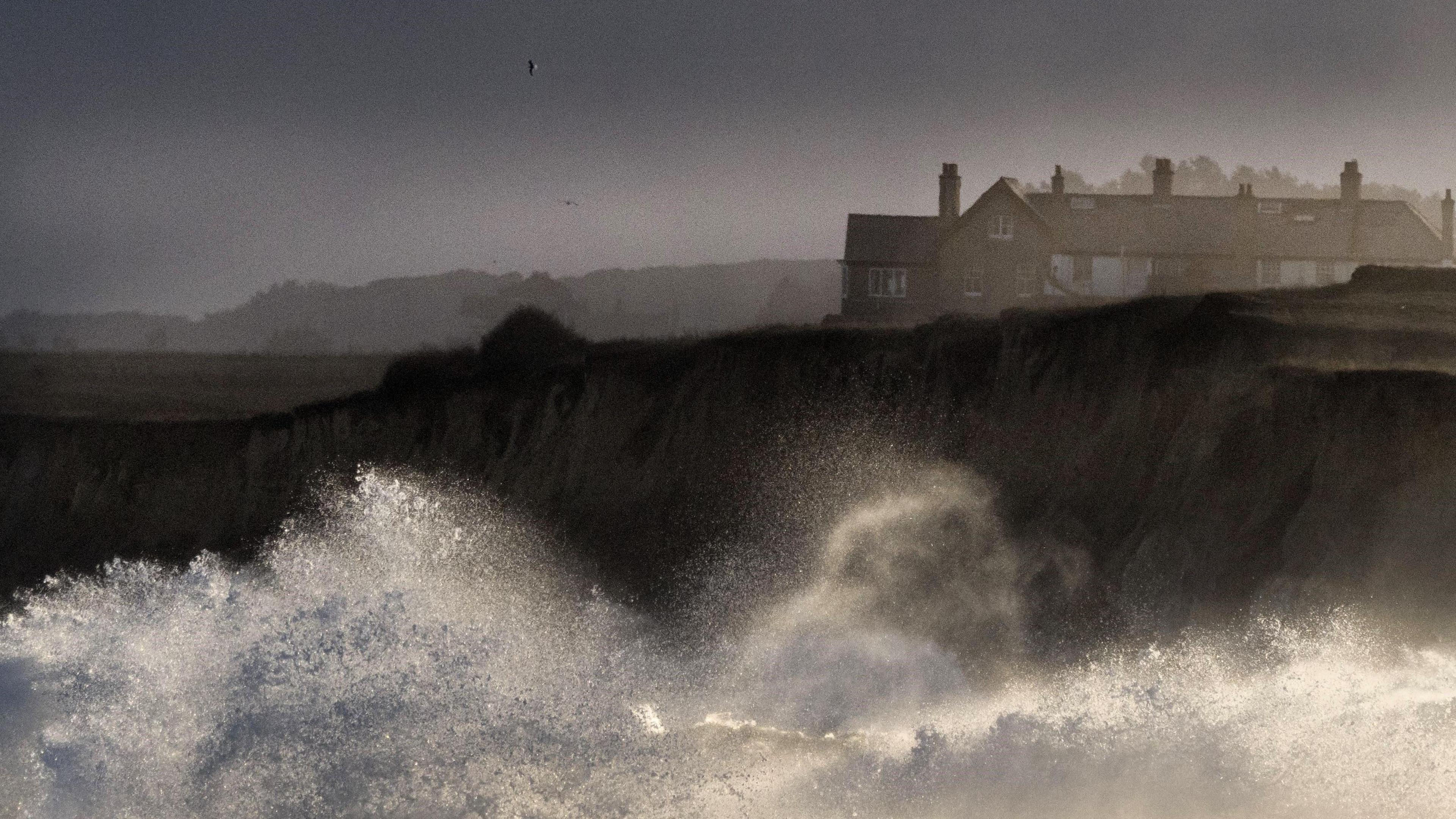 Large waves crashing on sandy cliffs with cliff-top homes in the background