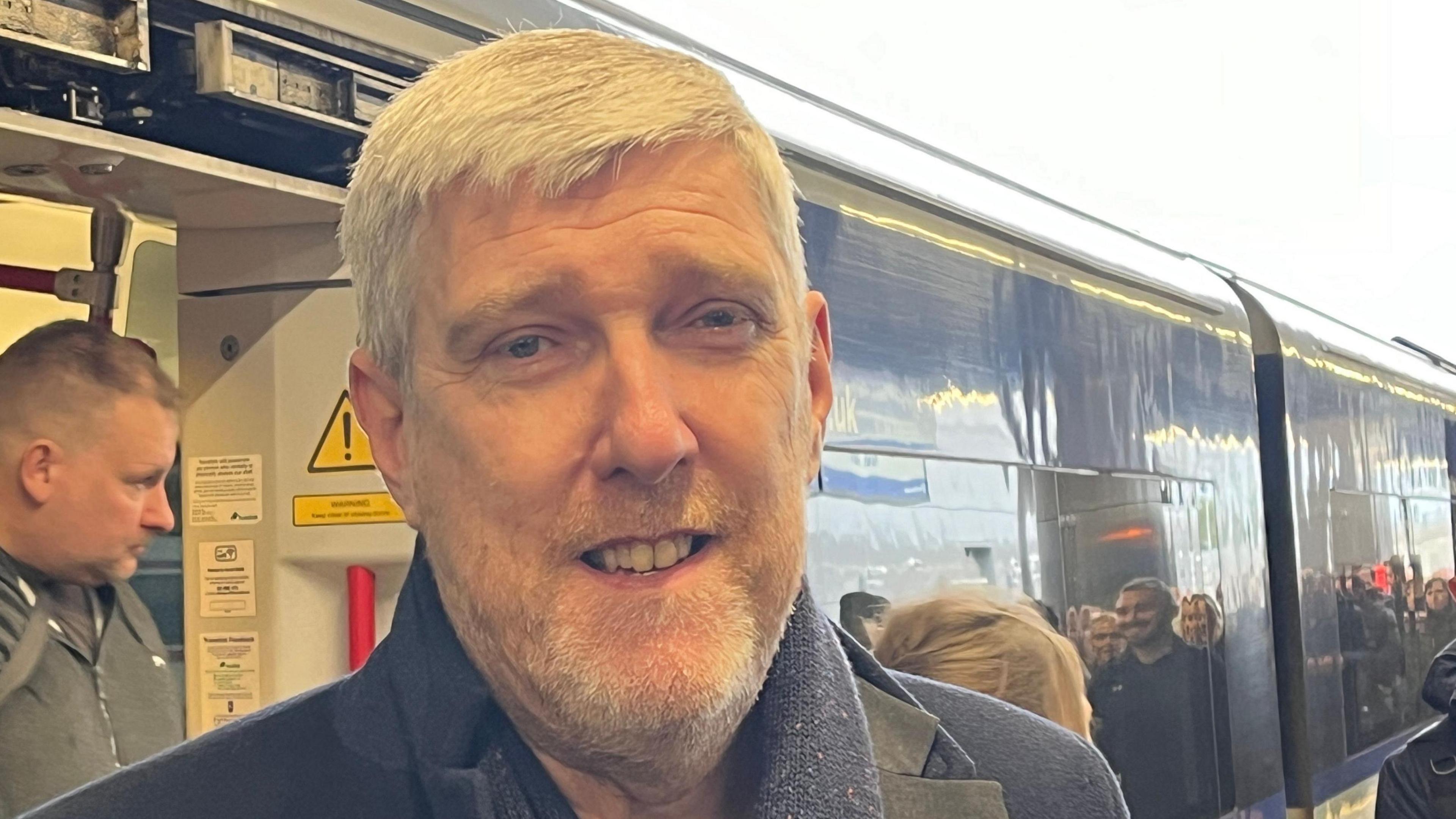 John O'Dowd with white short hair and beard, smiling at the camera in front of a train with passengers coming off - he is wearing a navy coat and scarf 