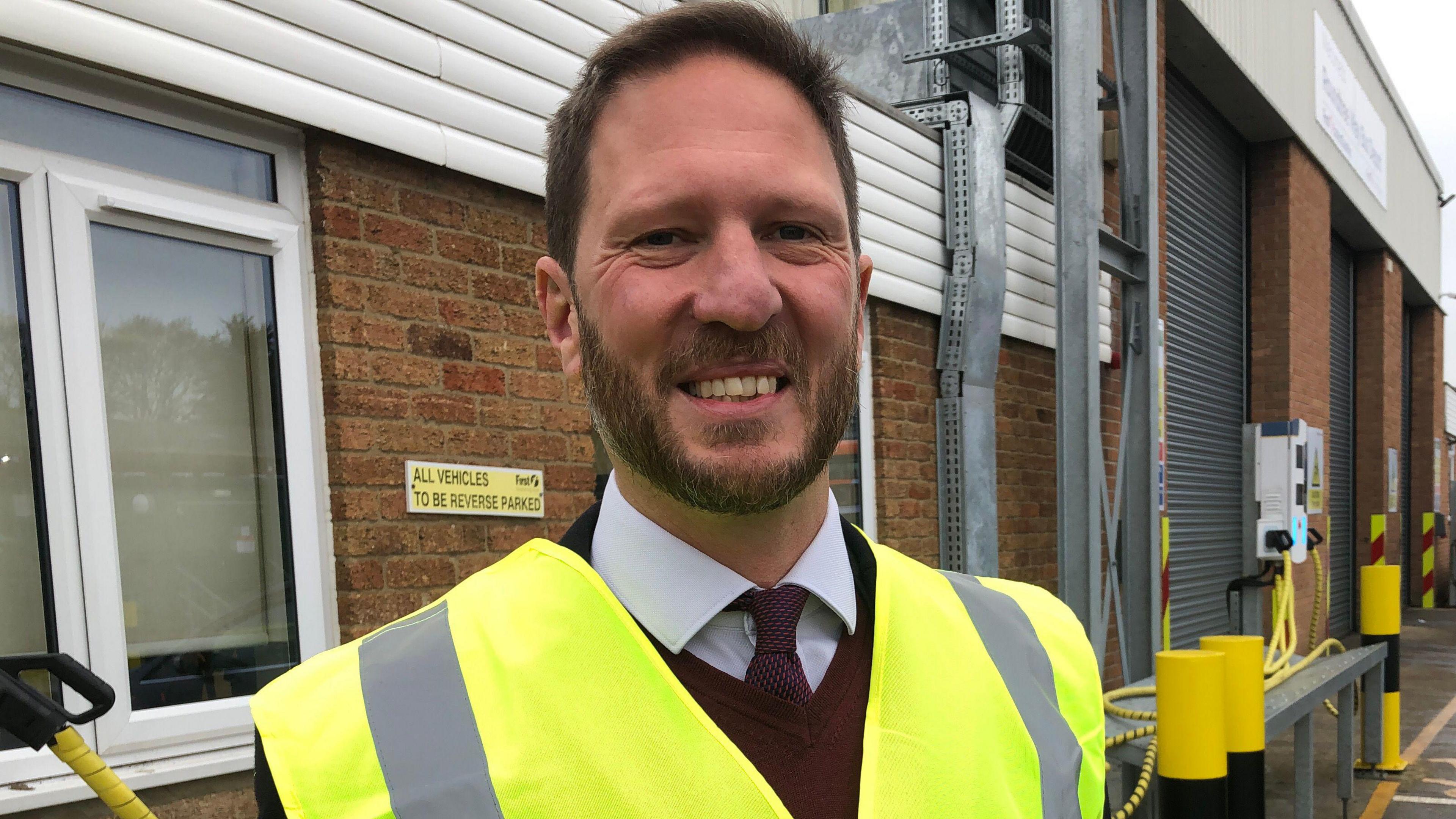 James McCluskey, a man in a white shirt, a red tie and red jumper. He is wearing a fluorescent hi-vis jacket and is looking into the camera and smiling. 