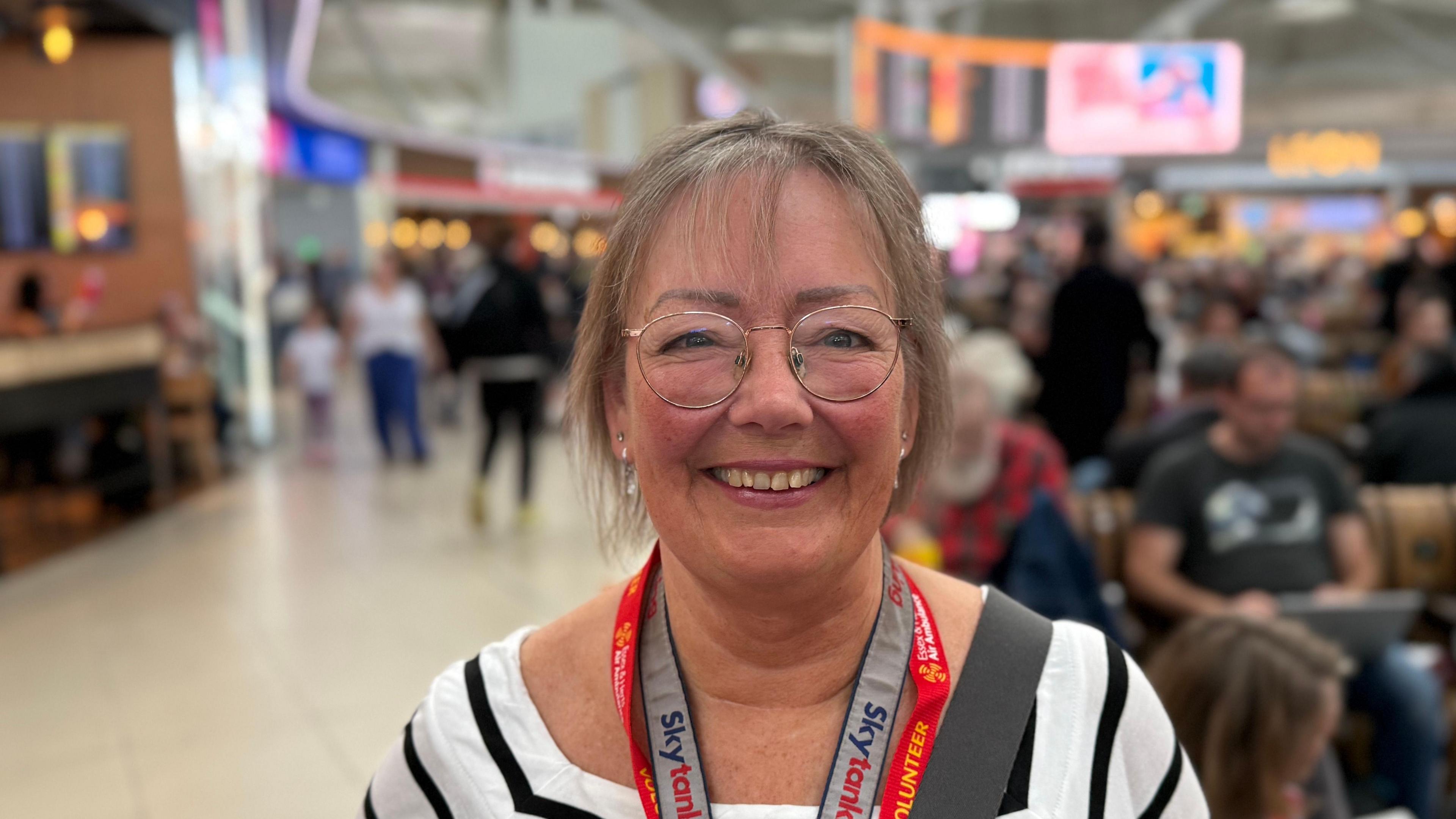 Alison is standing inside Stansted airport wearing a white top with black stripes across it. She is also wearing glasses and lanyards. In the background there are passengers in a busy terminal.