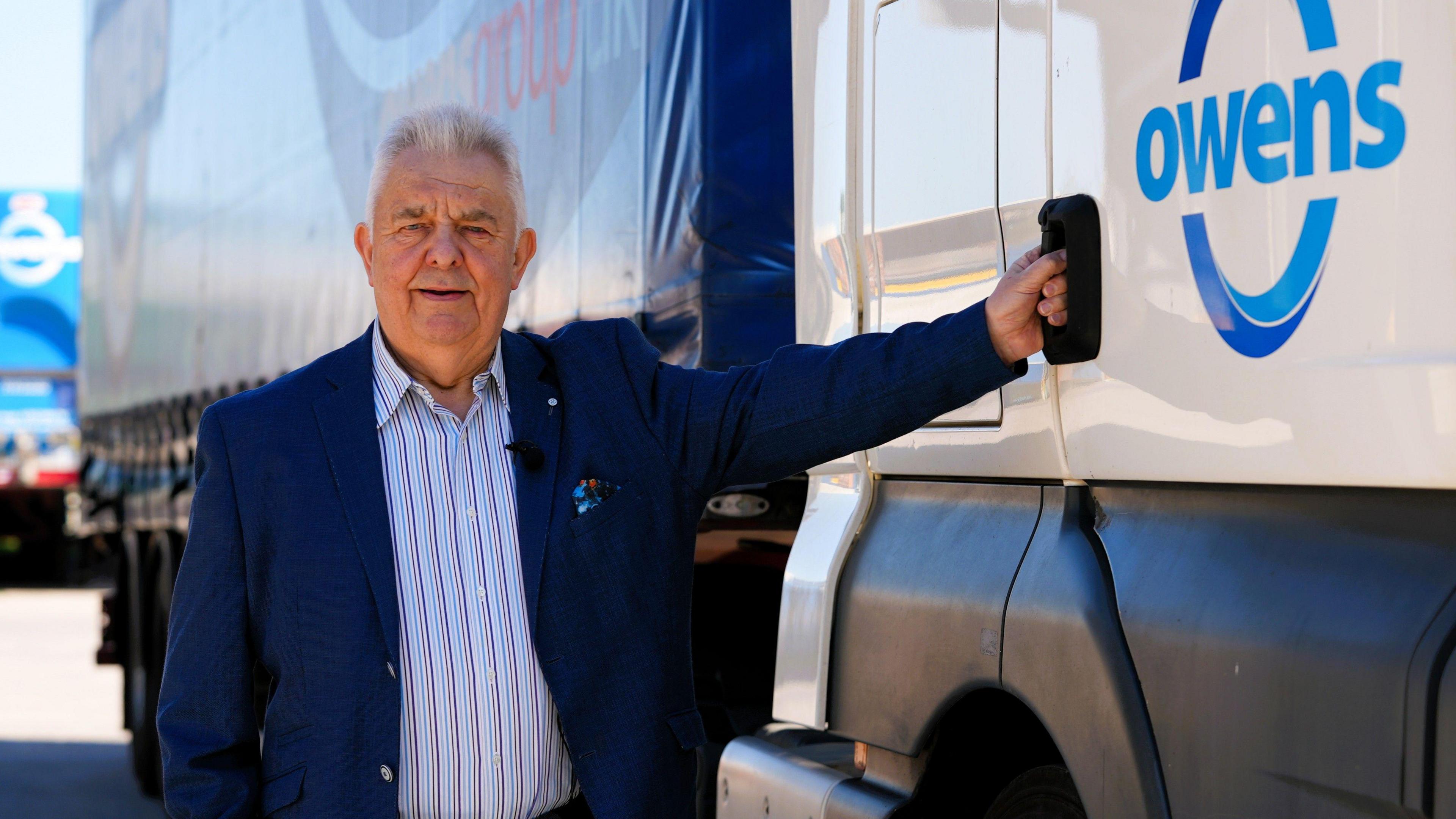 Dyn mewn siaced glas tywyll a chrys golau yn sefyll wrth ymyl lori gywn a glas o gwmni Owens