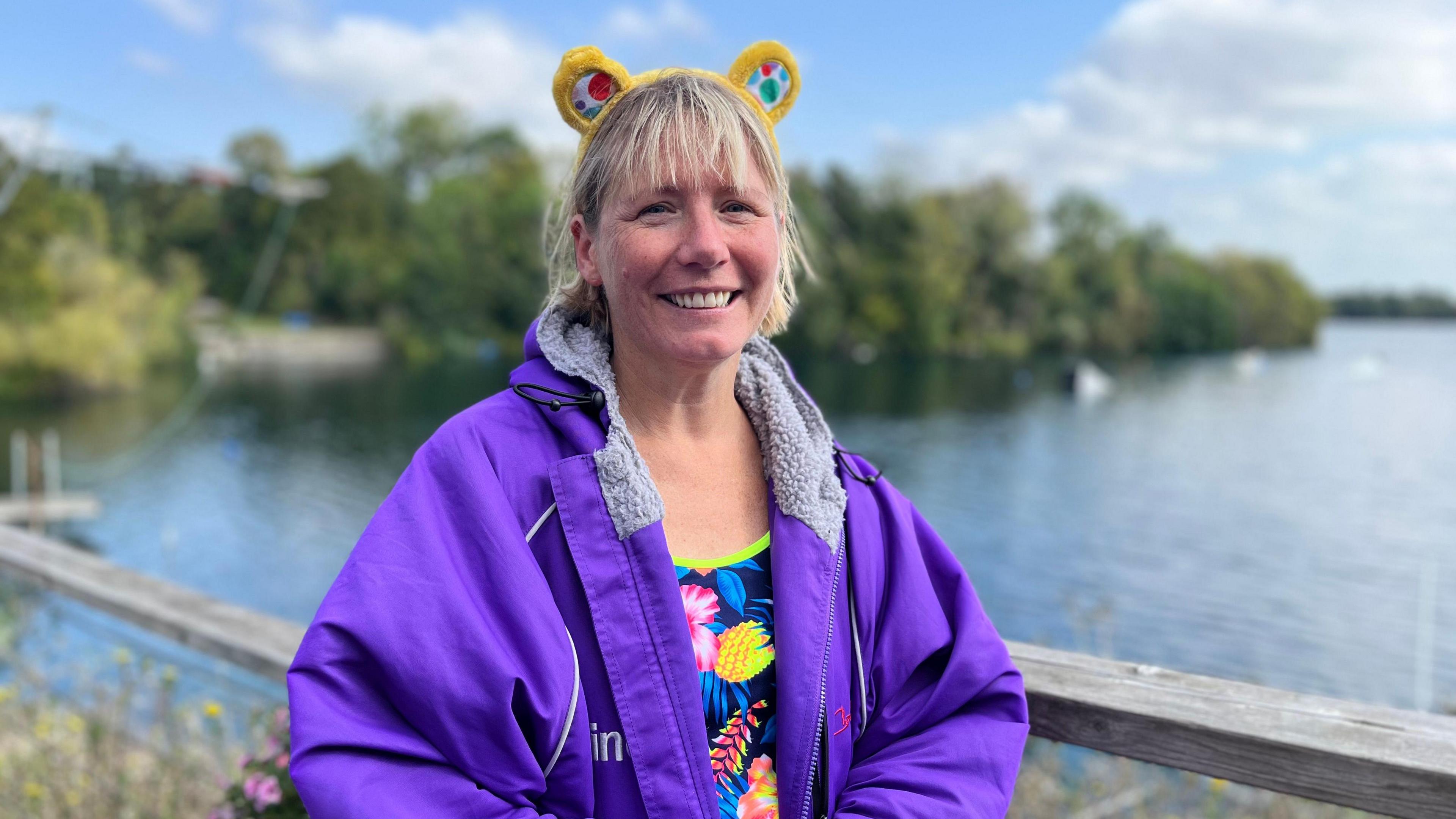 A woman wearing a purple waterproof coat over a multicoloured swimming costume. She's wearing a pair of yellow Pudsey Bear ears and smiling at the camera with a lake in the background.