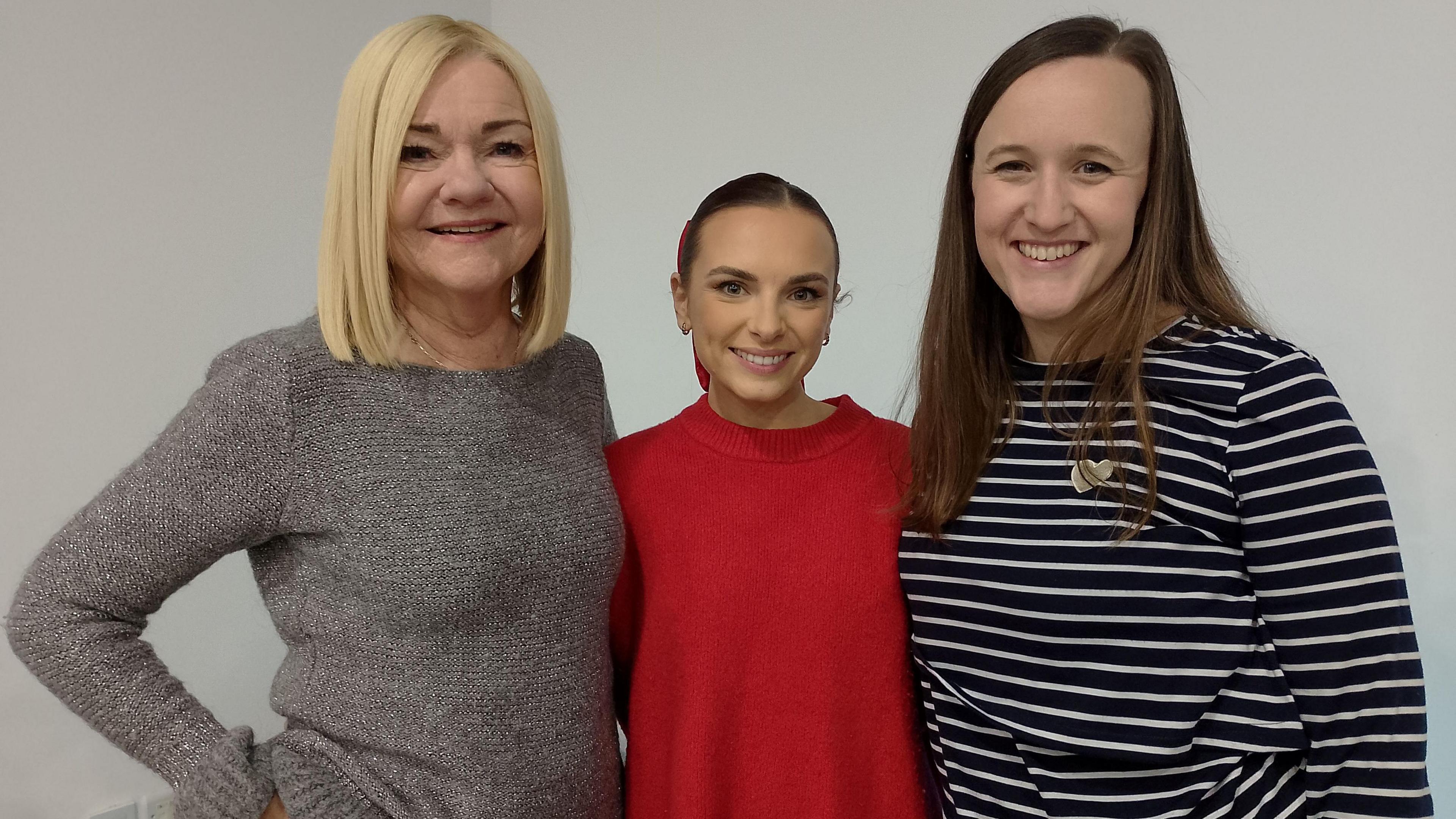 Three women stand together. Paula Gibson has shoulder-length blond hair and is wearing a grey jumper. Amber Harrison is wearing a red jumper and has brown hair pulled back. Christelle Lees has long brown hair and is wearing a black and white stripped top.