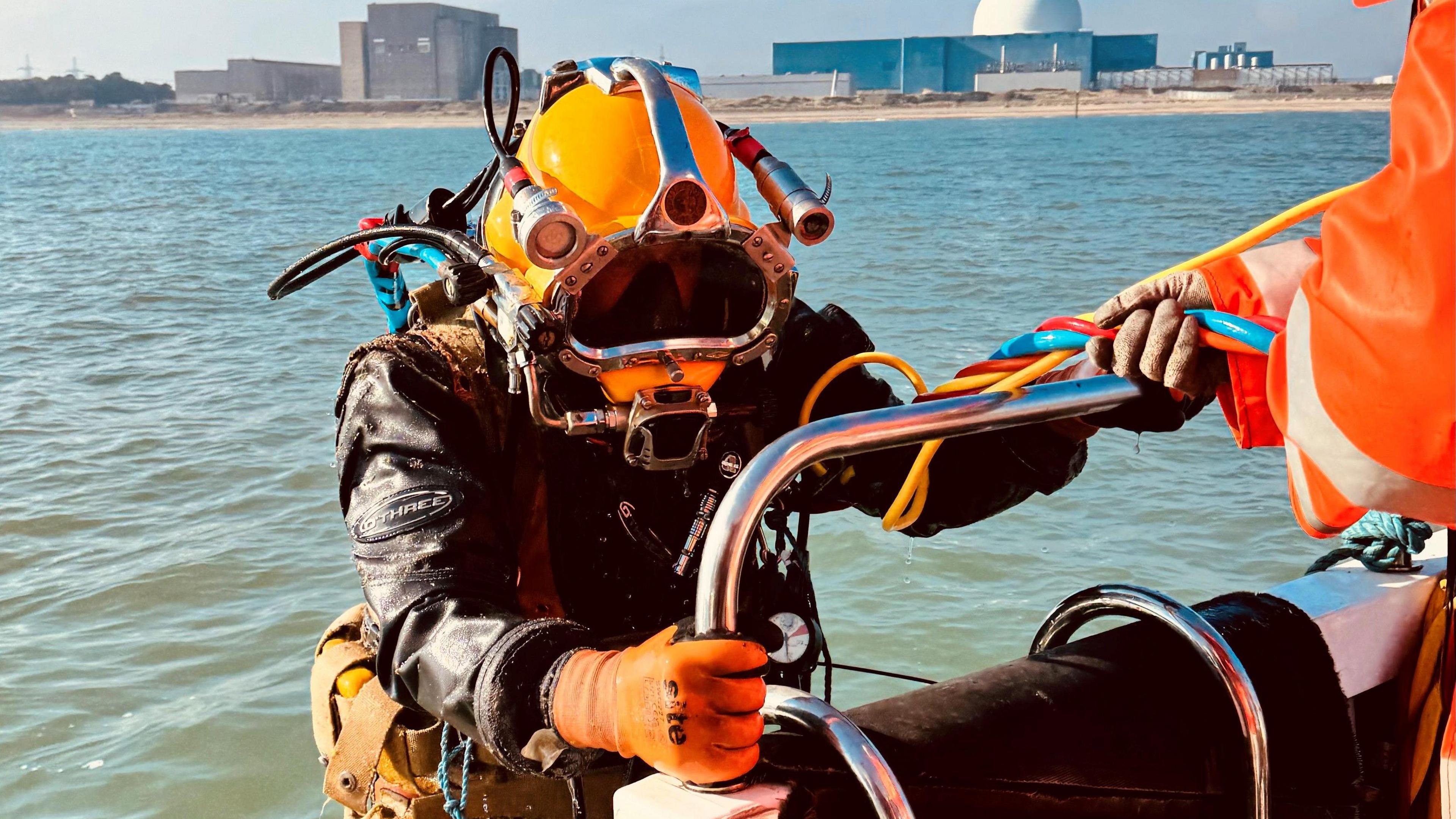 A diver holding onto the side of a platoon floating in the sea. The diver is wearing a diving suit and has there is someone in a high-visibility orange jacket helping him. 