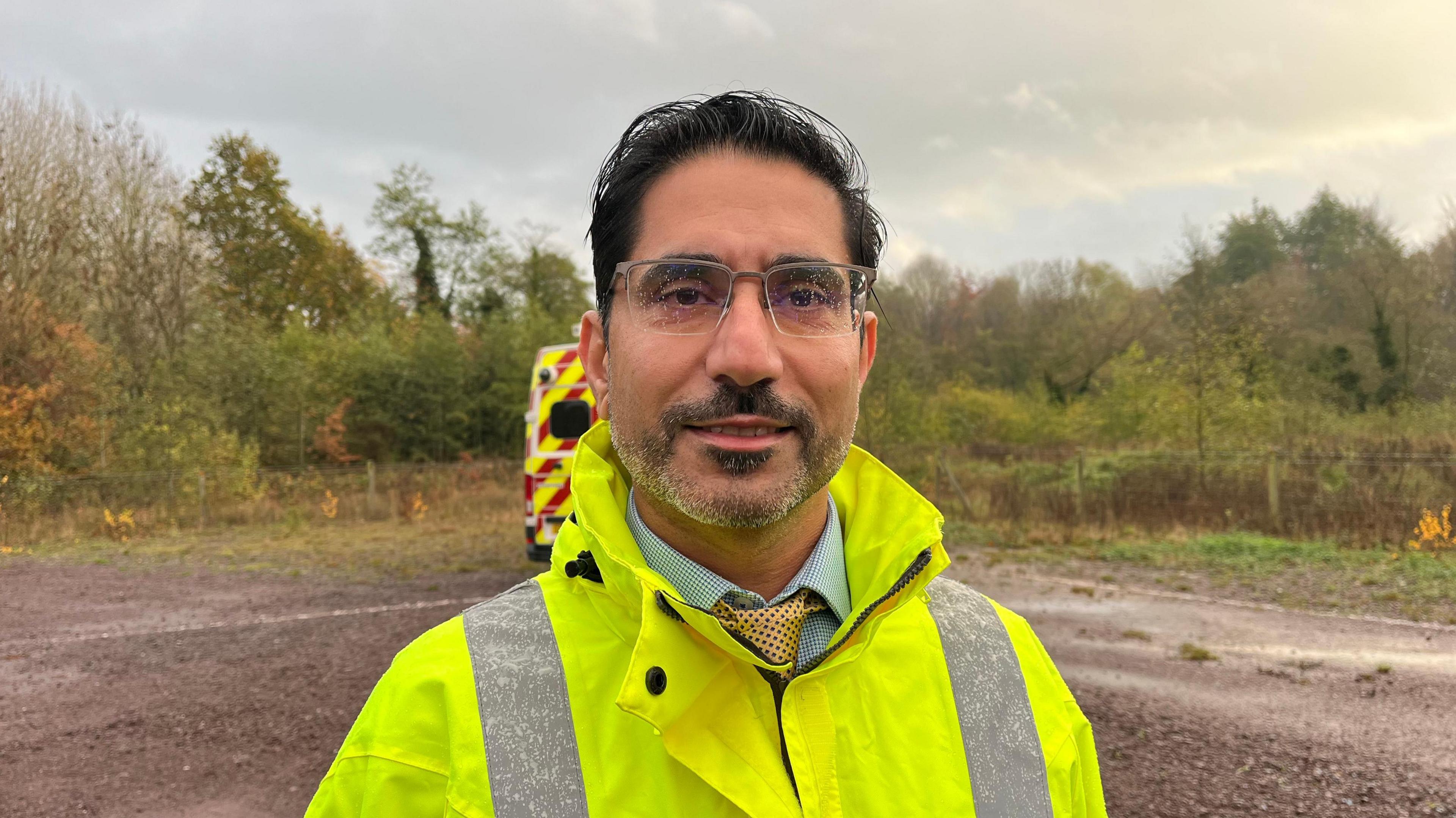 Amitabh pictured smiling slightly and looking into the camera. Her has dark hair, a short stubbly beard and is wearing glasses. He's wearing a bright yellow hi-vis raincoat, and has a light blue shirt and yellow tie on underneath. He's stood in a muddy, gravel car park that has trees and shrubbery along the edges. The trees' leaves are green and orange, and the sky is grey and overcast. 