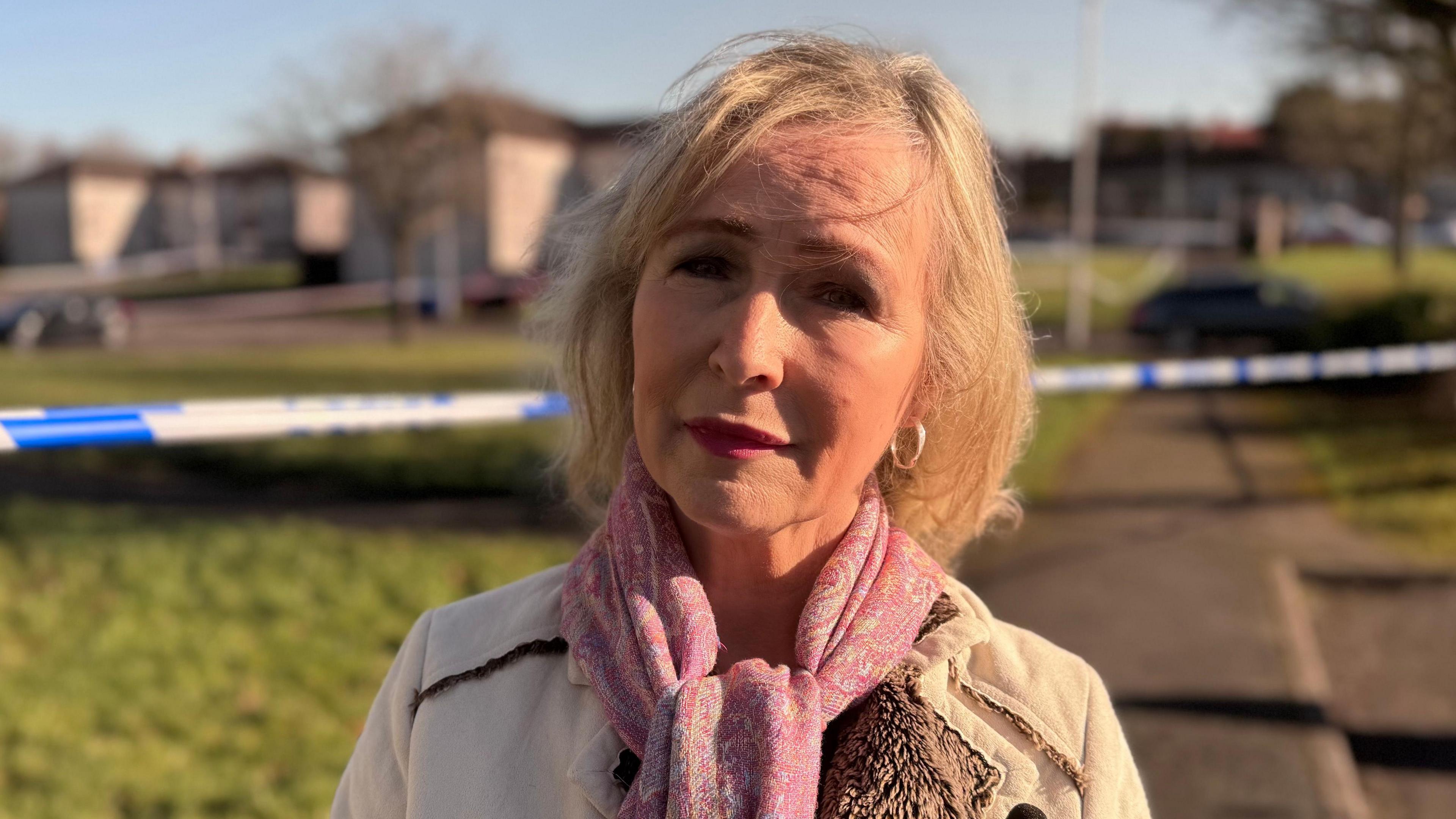 Deirdre has blond shoulder length hair, gold hoop earrings and a pink scarf and cream coat. 
She is standing in front of of a police cordon. The houses behind are blurred out. 