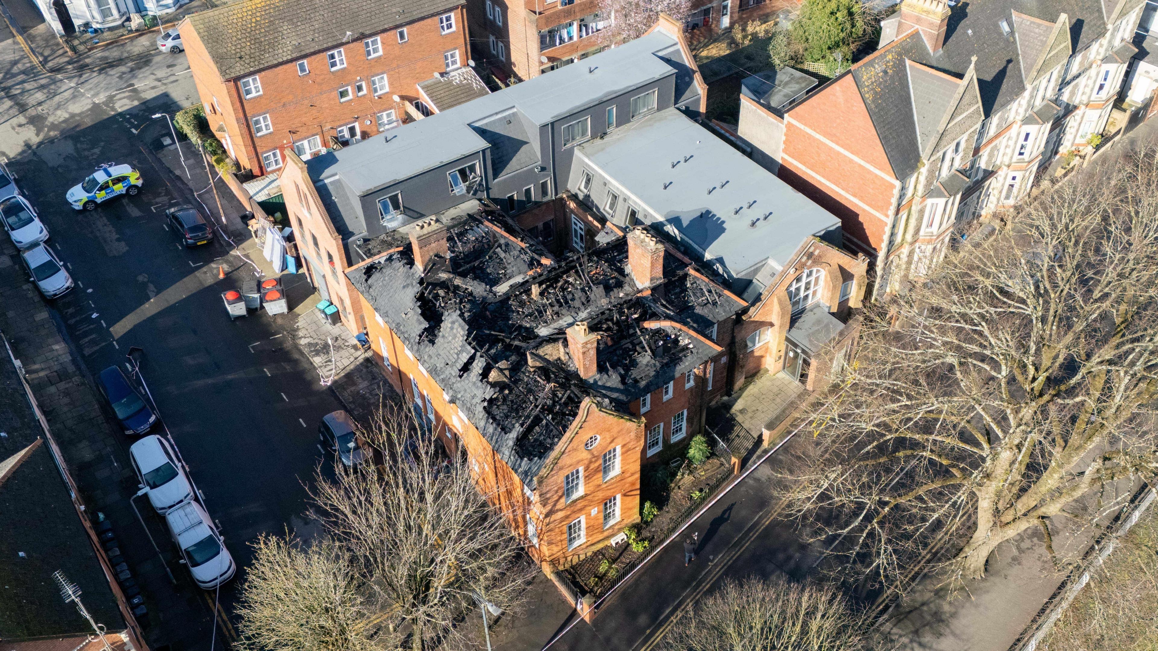 Aerial view of the large property from a greater distance showing the damaged roof and surrounding houses. The roof is completely collapsed with only burnt blackened timbers visible.