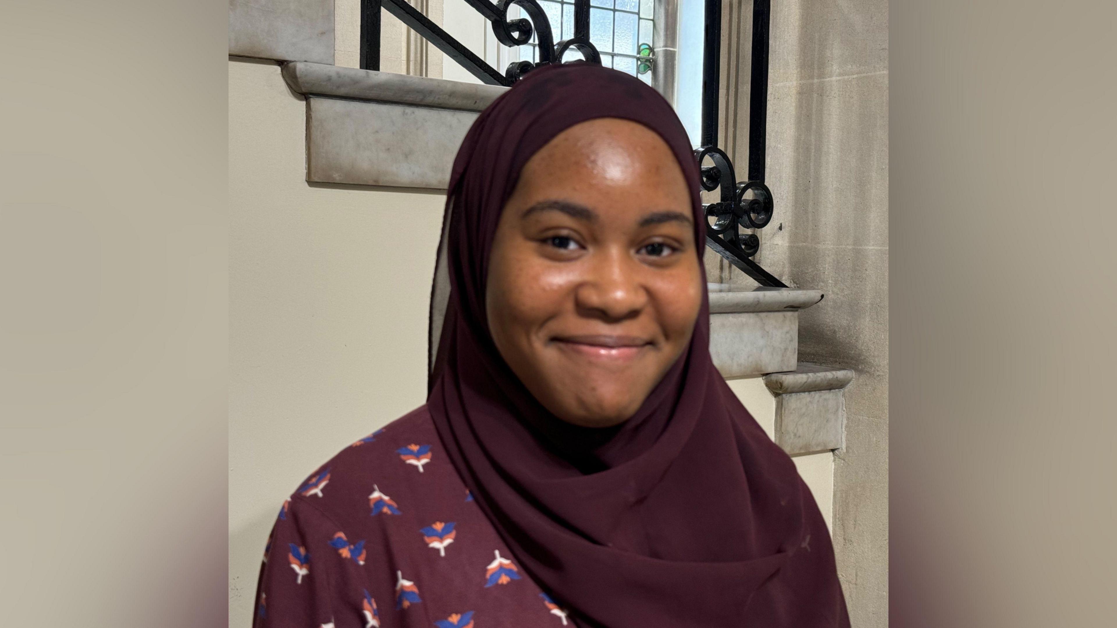 Picture of Ayyat Adigun smiling and wearing a headscarf in front of stone steps and a stained glass window.