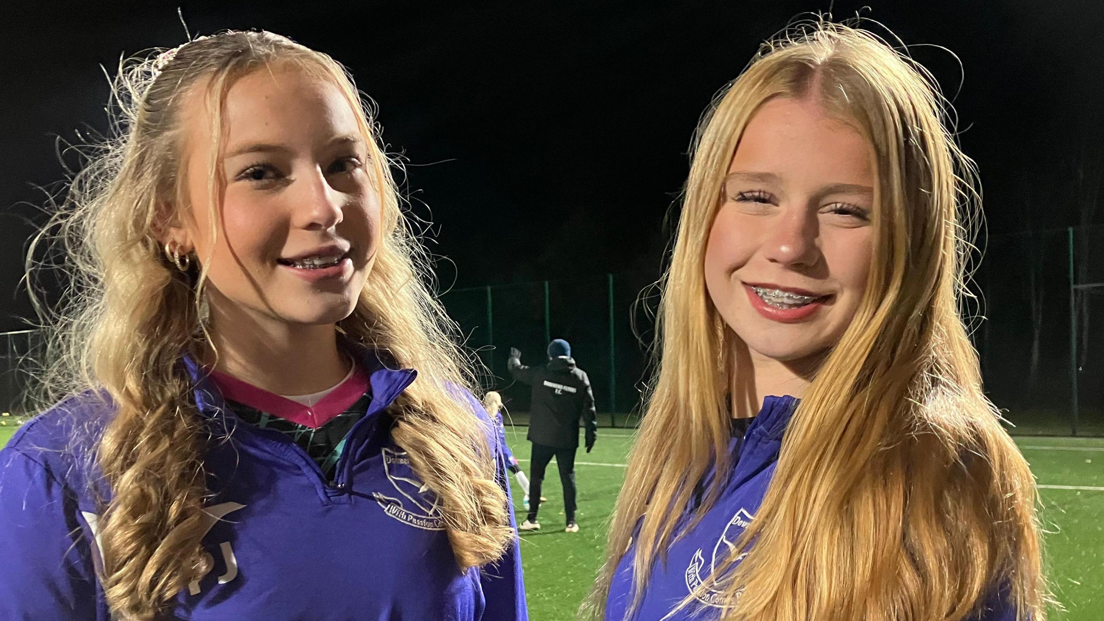 Two young girls are smiling for the camera. They are both wearing purple zip up jumpers with their football club branding on them in white. They are standing on a football pitch which is lit up with flood lights. They both have long blonde hair and braces.