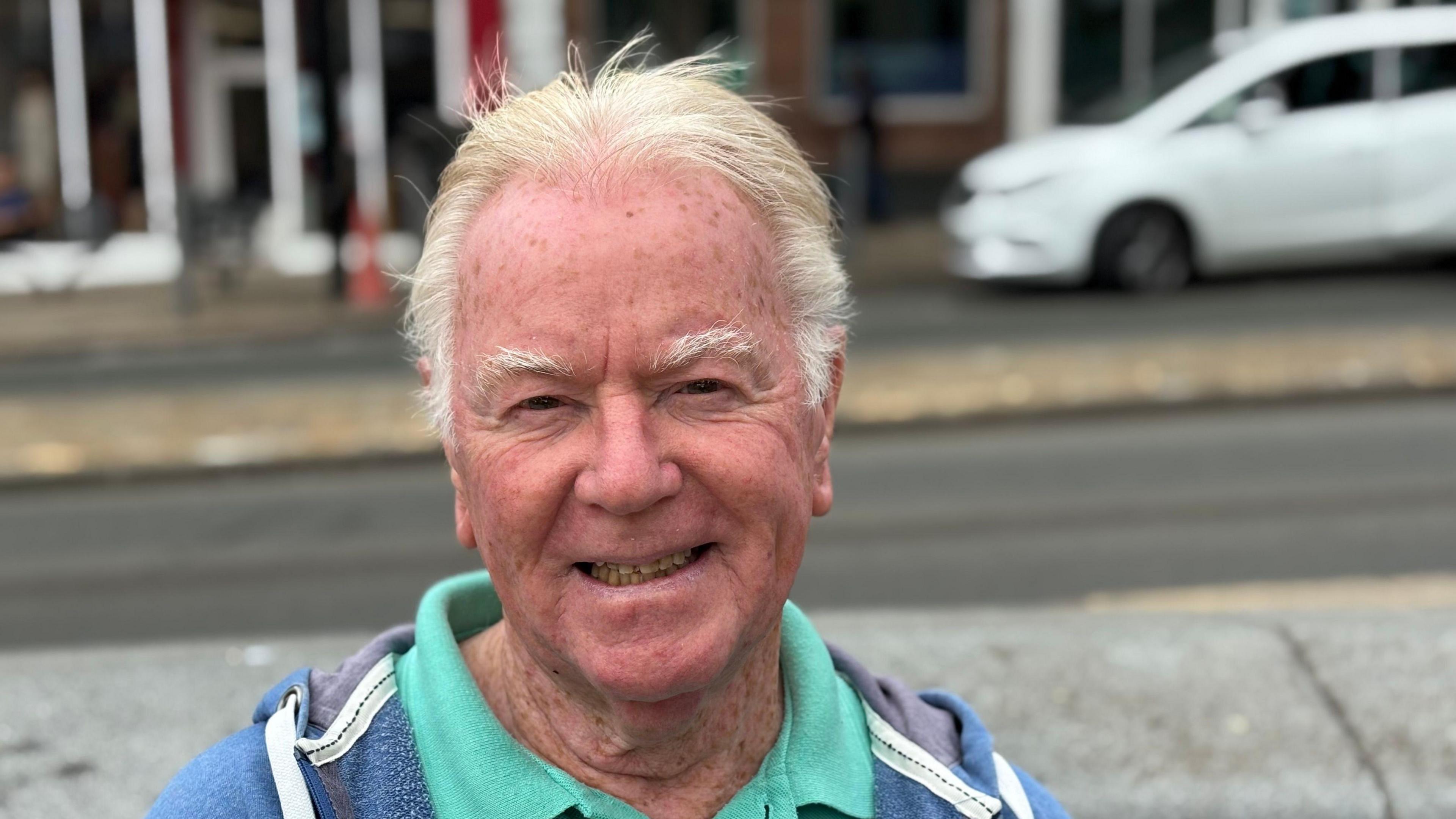 Man with white hair and wearing a turquoise polo shirt and blue hoodie is sitting on a bench in Wolverhampton City centre smiling