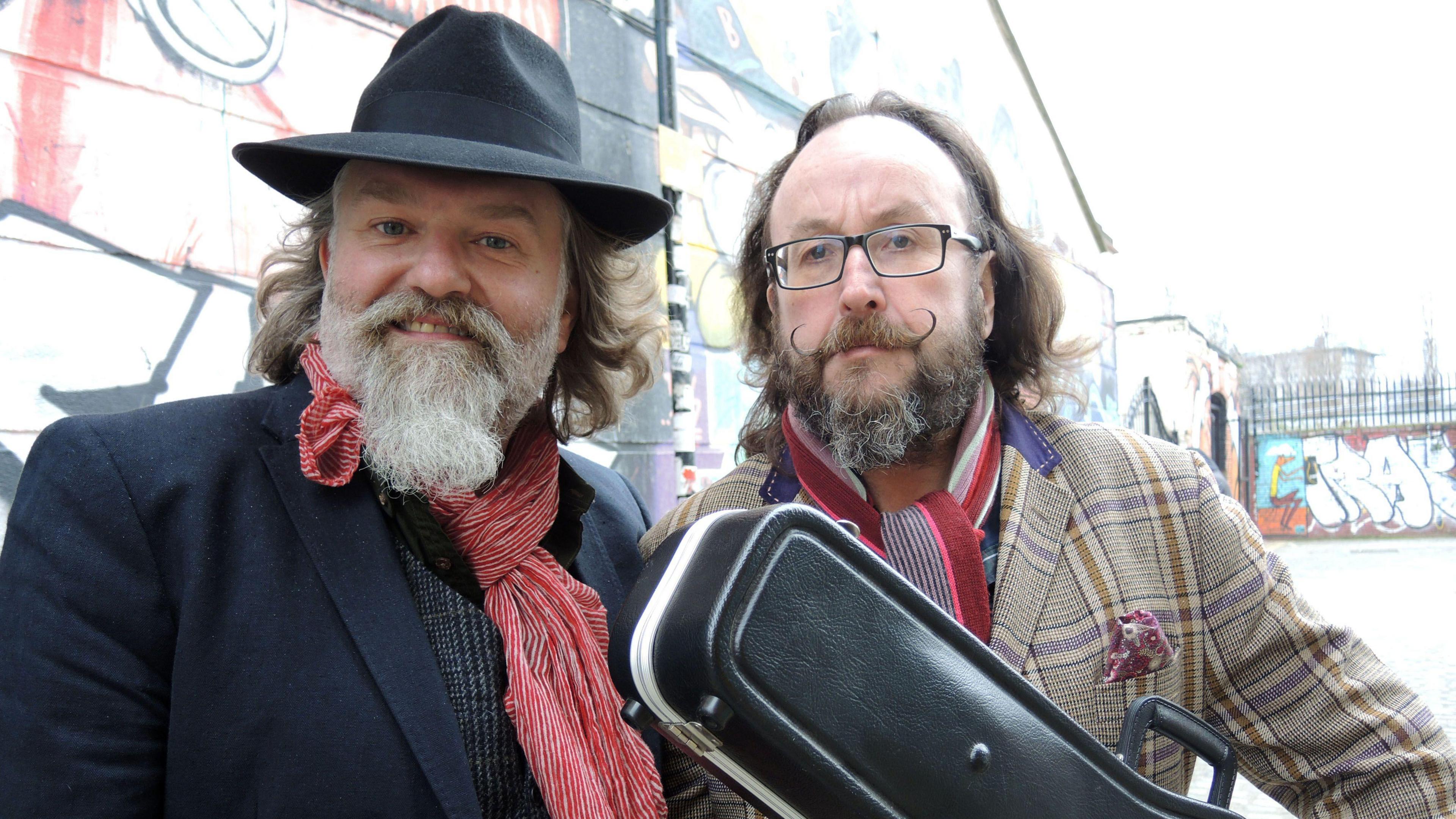 Si King and Dave Myers pose for the camera during the filming of one of their shows. King is wearing a black hat, and Myers is holding a guitar case.