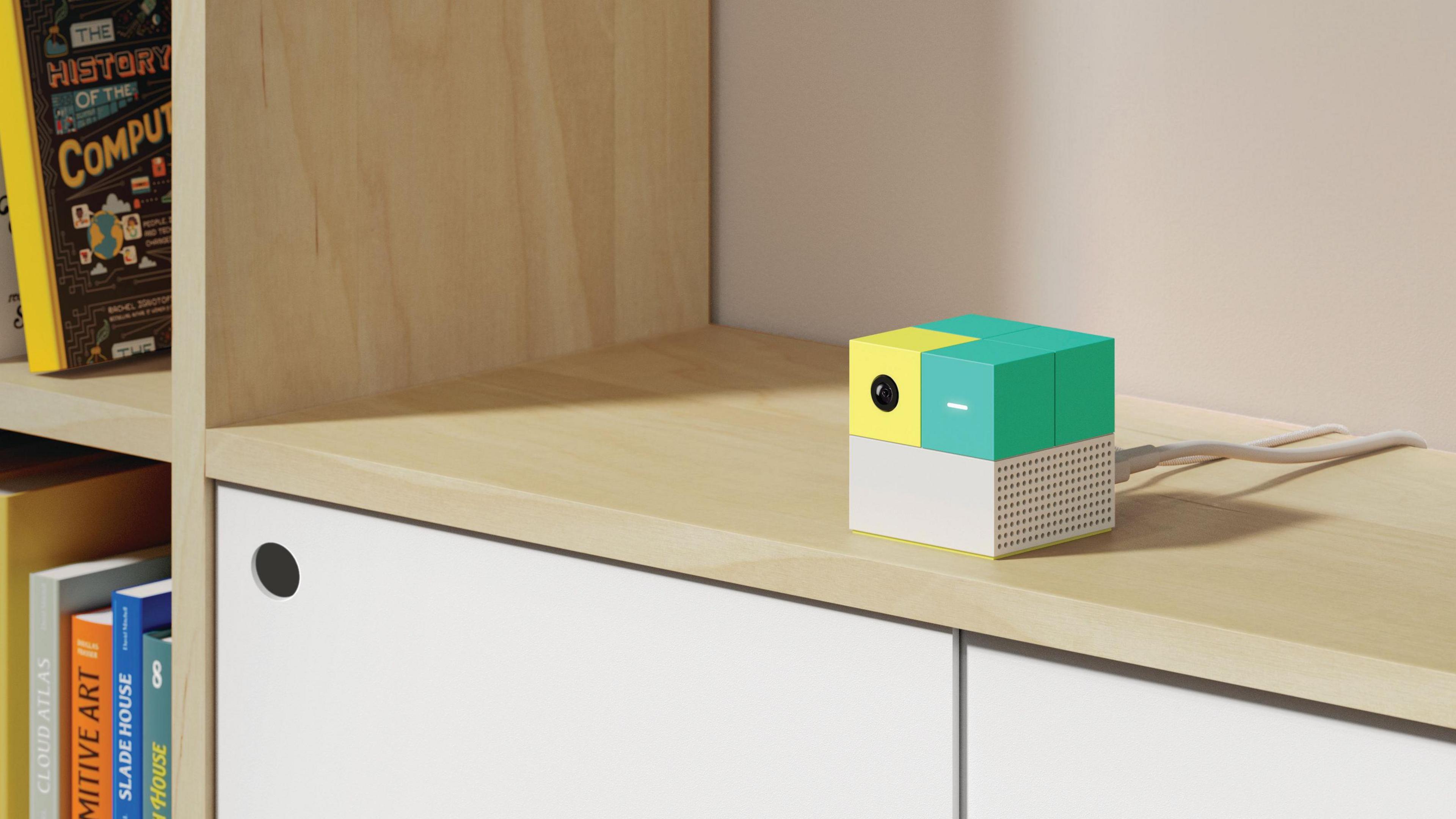 A close up on the Nex Playground console, a small green yellow and white cube on a surface, with white cables plugged into it, next to a bookshelf space