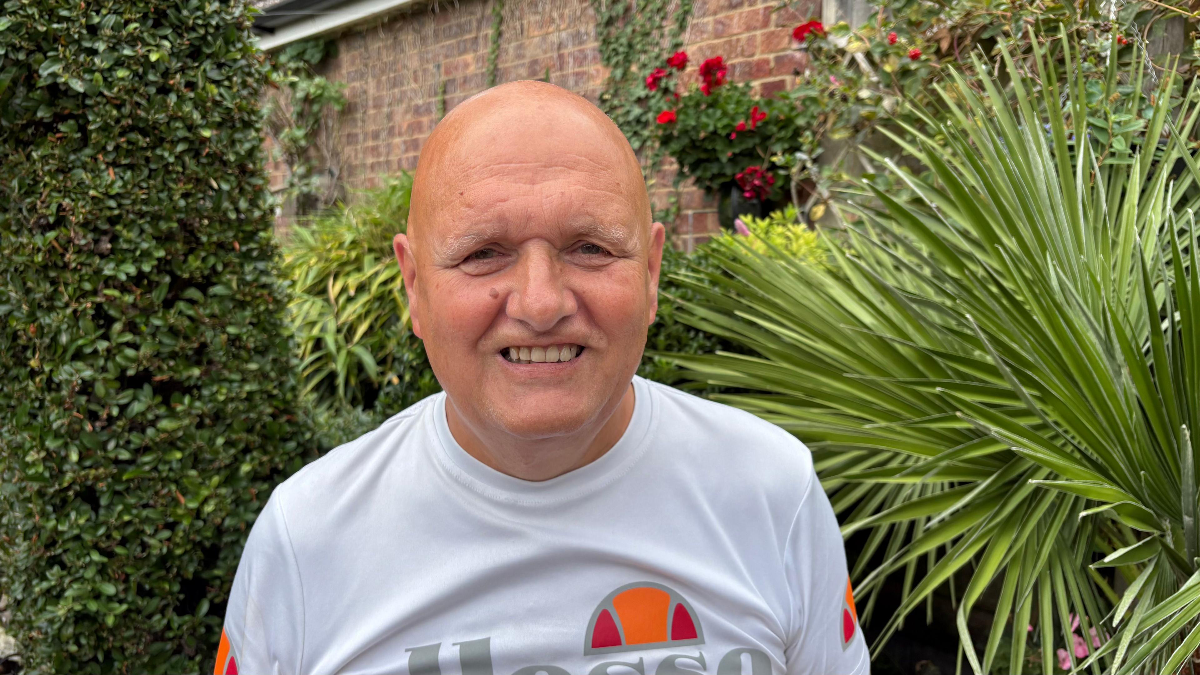 Rob with very short hair looking at the camera and wearing a white T-shirt with red and orange semi-circular logo. He is standing in front of ferns and trees in a garden.