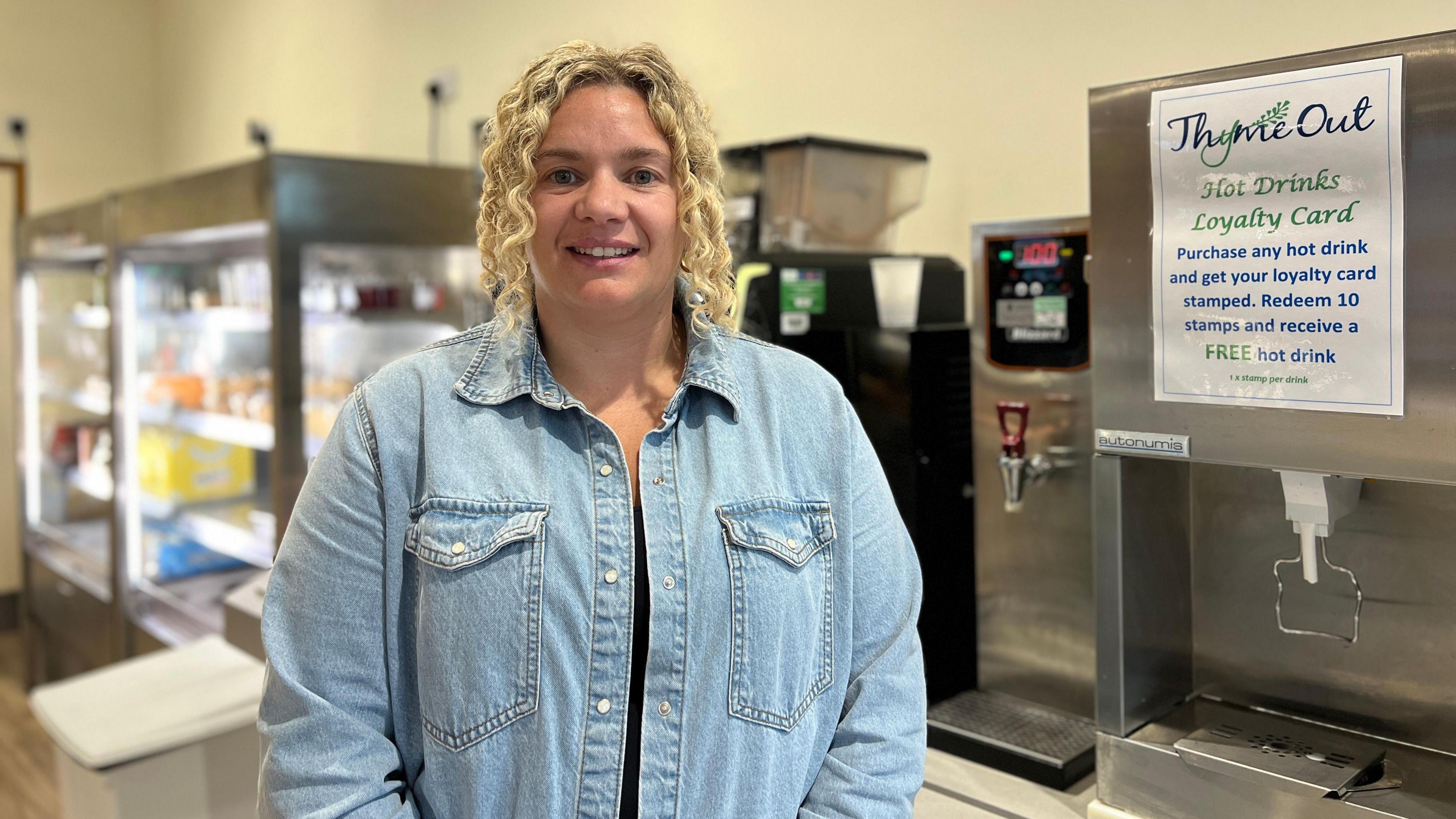 Ella smiles at the camera as she holds her hands in front of her. She is wearing a light blue denim jacket and to her left is a hot drinks machine which has the cafe's name on a sign on it. Behind her to her right is a fridge unit with drinks and sandwiches inside.