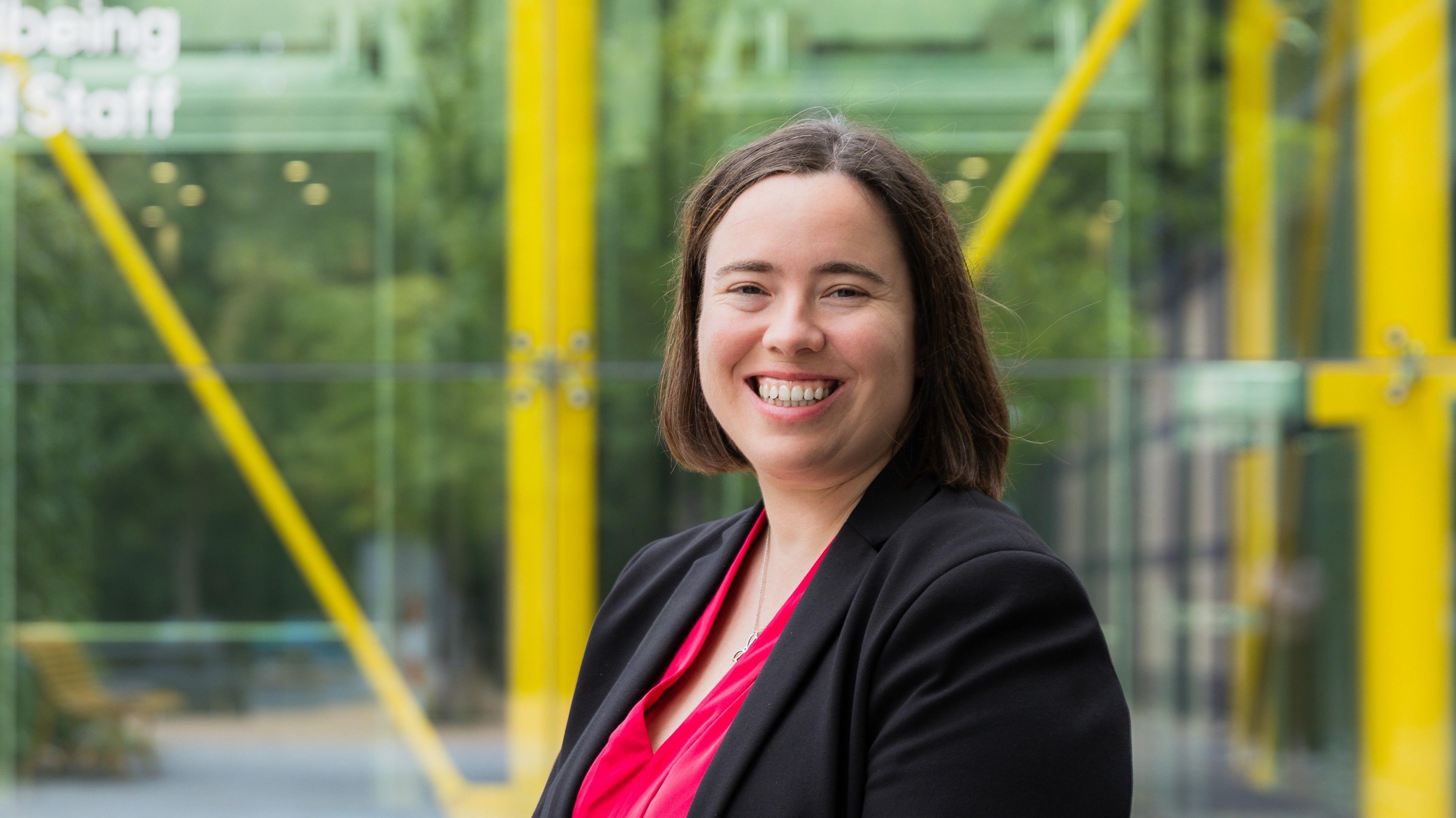 Dr Georgina Marsh  looks slightly over her shoulder to look at the camera and smile. She stands inside a glass building with yellow structural poles. She has short brown hair and wears a black blazer with a bright pink blouse underneath. 