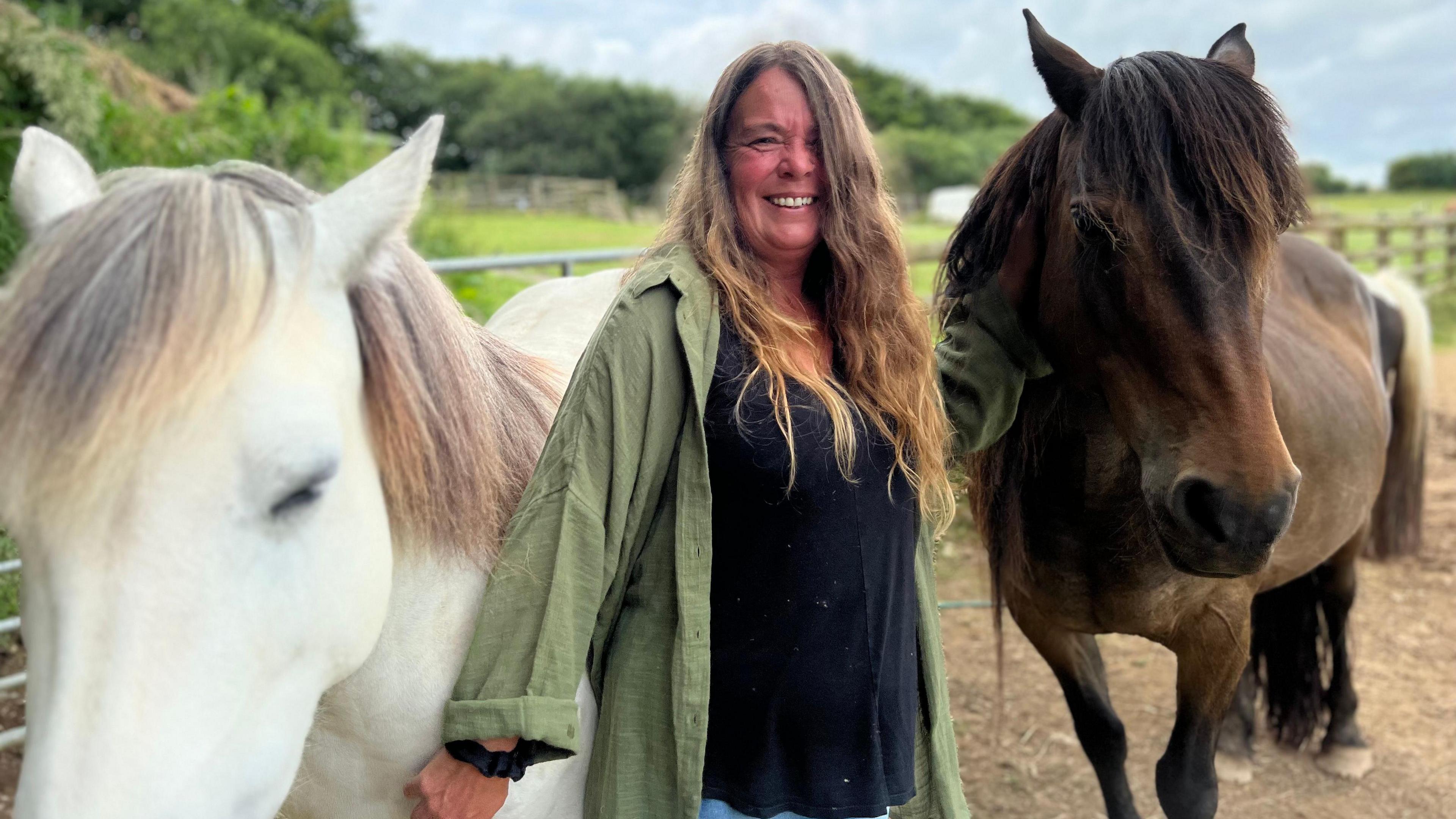 A woman with long hair smiles as she is flanked by a white horse and a brown horse