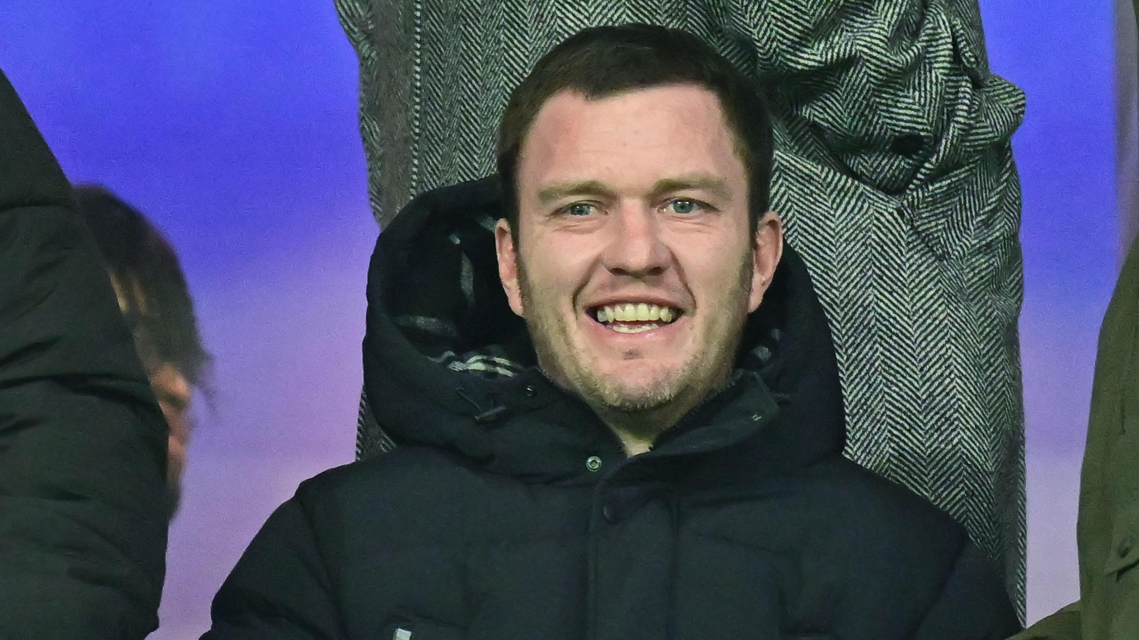 Craig Gardner sits in the directors' box in a dark puffer coat. He is smiling and behind him someone is standing up in a black and white tweed overcoat.