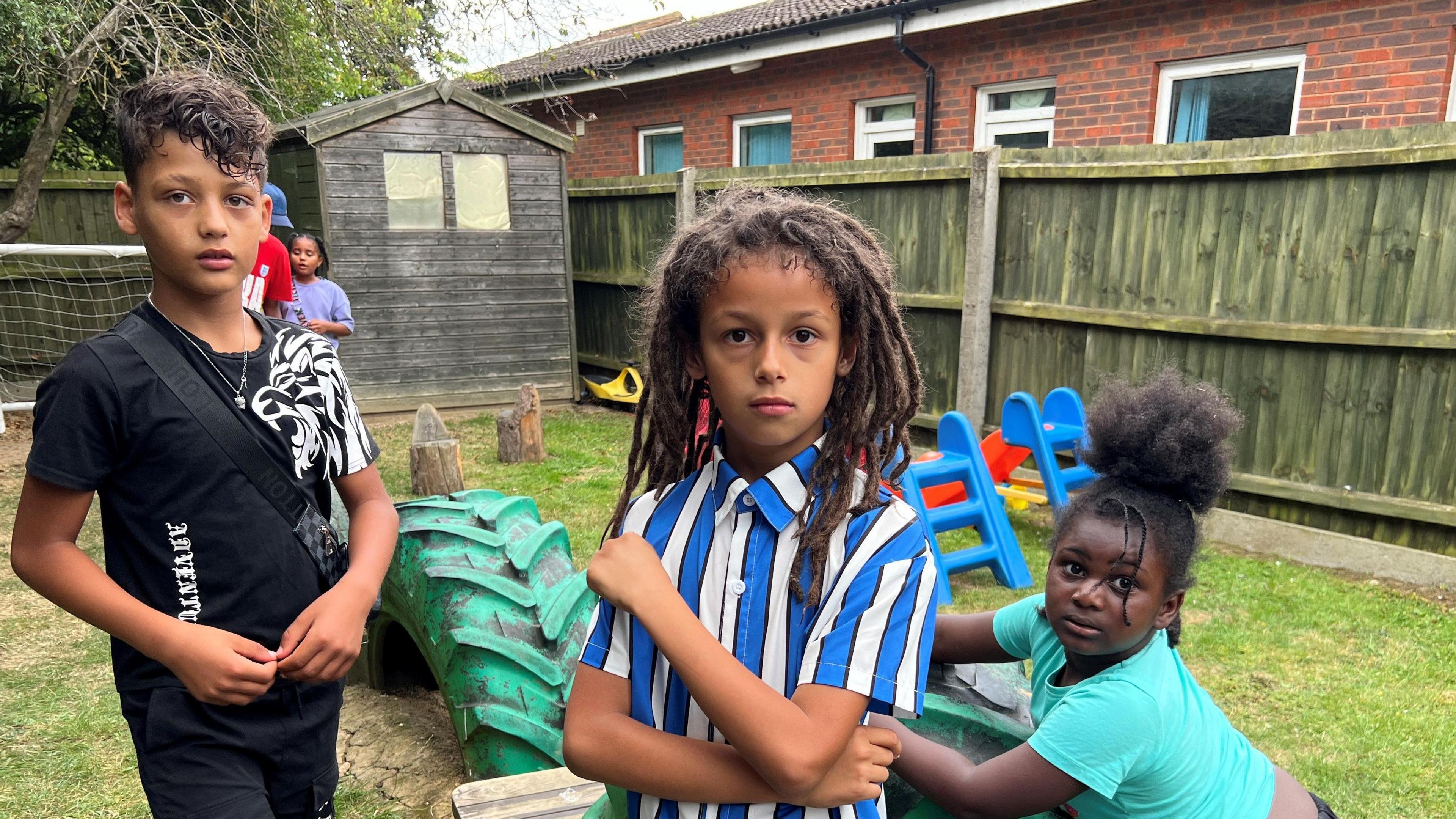 Children enjoying the Saturday school garden. In the foreground, a child with a short back and sides hair cut, with long waves on top in a black t-shirt with a silver lion on his shoulder. Next to him is a boy with long dreds wearing a chunky, blue, white and black striped shirt. On the right is a girl with a pineapple hairdo with plaited tendrils at the front, wearing a green t-shirt. Behind them are a green tyre, slide and other garden play equipment and other children