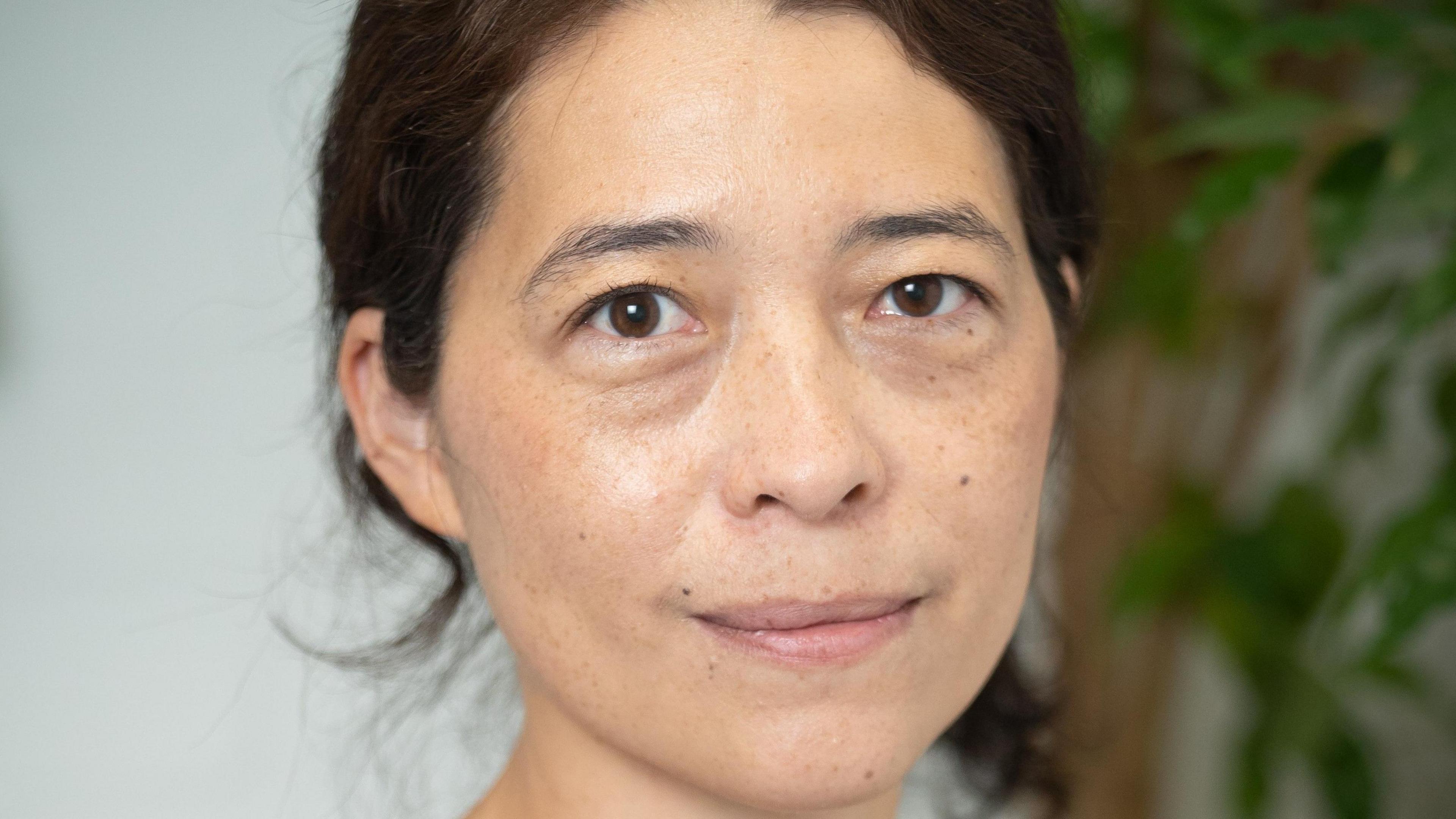 Headshot of Akiko Hart looking directly at the camera. Her eyes are brown, her black hair is tied behind her head and she has pursed lips. Over her left ear, there is an out-of-focus plant in the background.