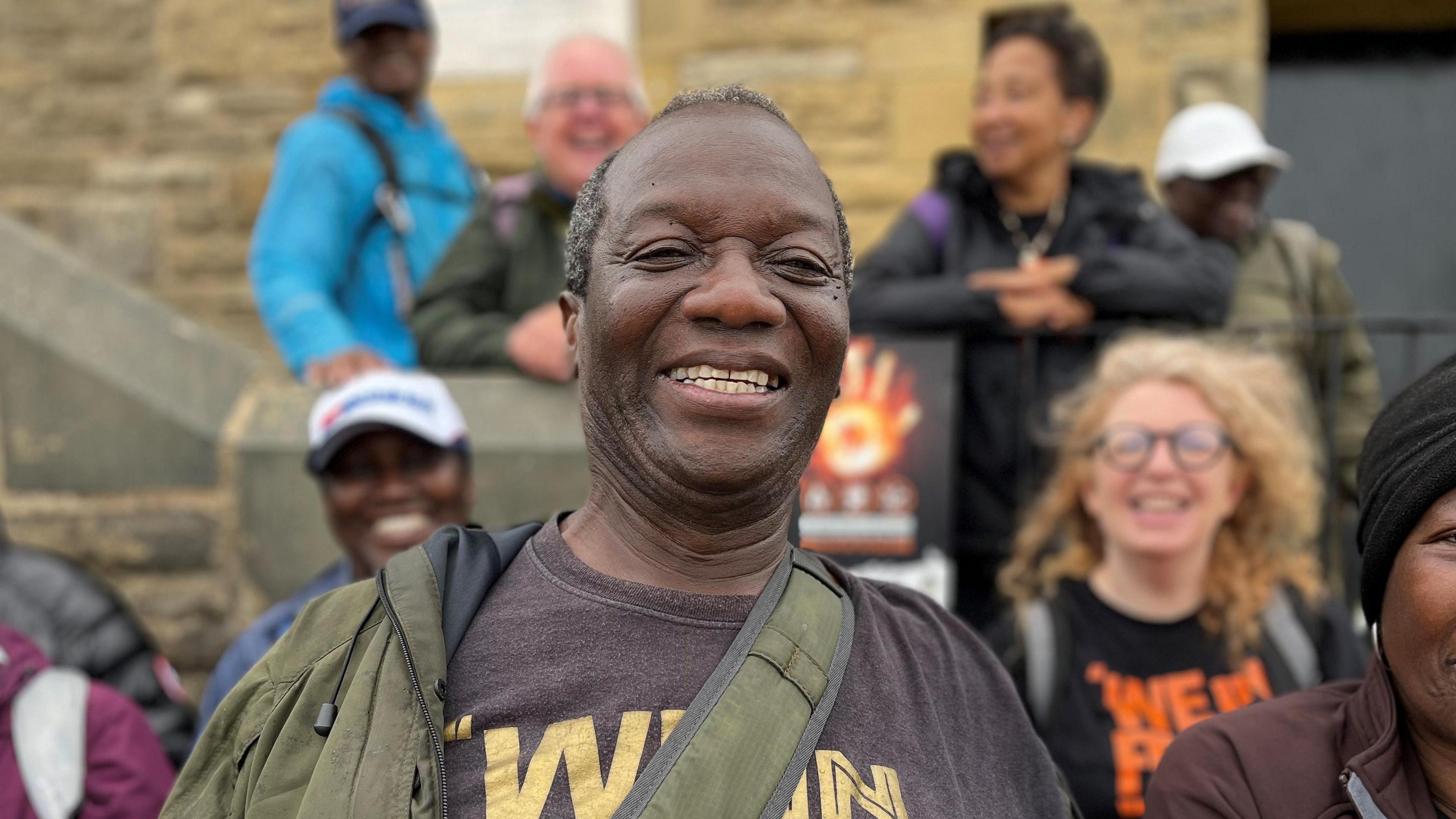 A West Indian man in his seventies smiles directly at the camera. He is wearing a grey t-shirt and a green waterproof, with a green cross body bag. Although the background is blurred, other people can be seen smiling at the camera too - a white woman with blonde curly hair and glasses, and a black woman with a white baseball cap.