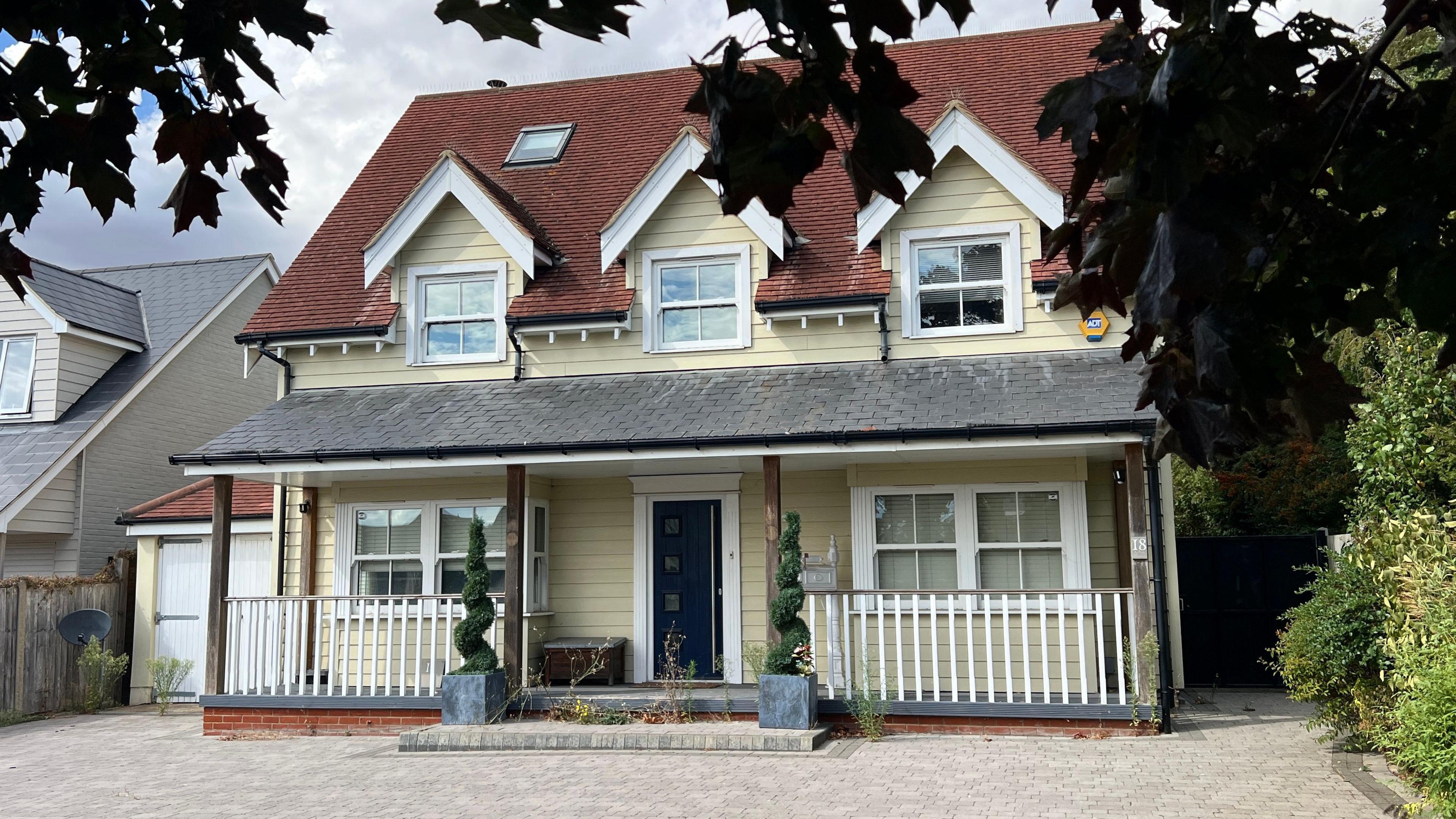 The front of a light yellow weatherboarded house of three storeys. It has a large driveway and two potted plants by the front door.