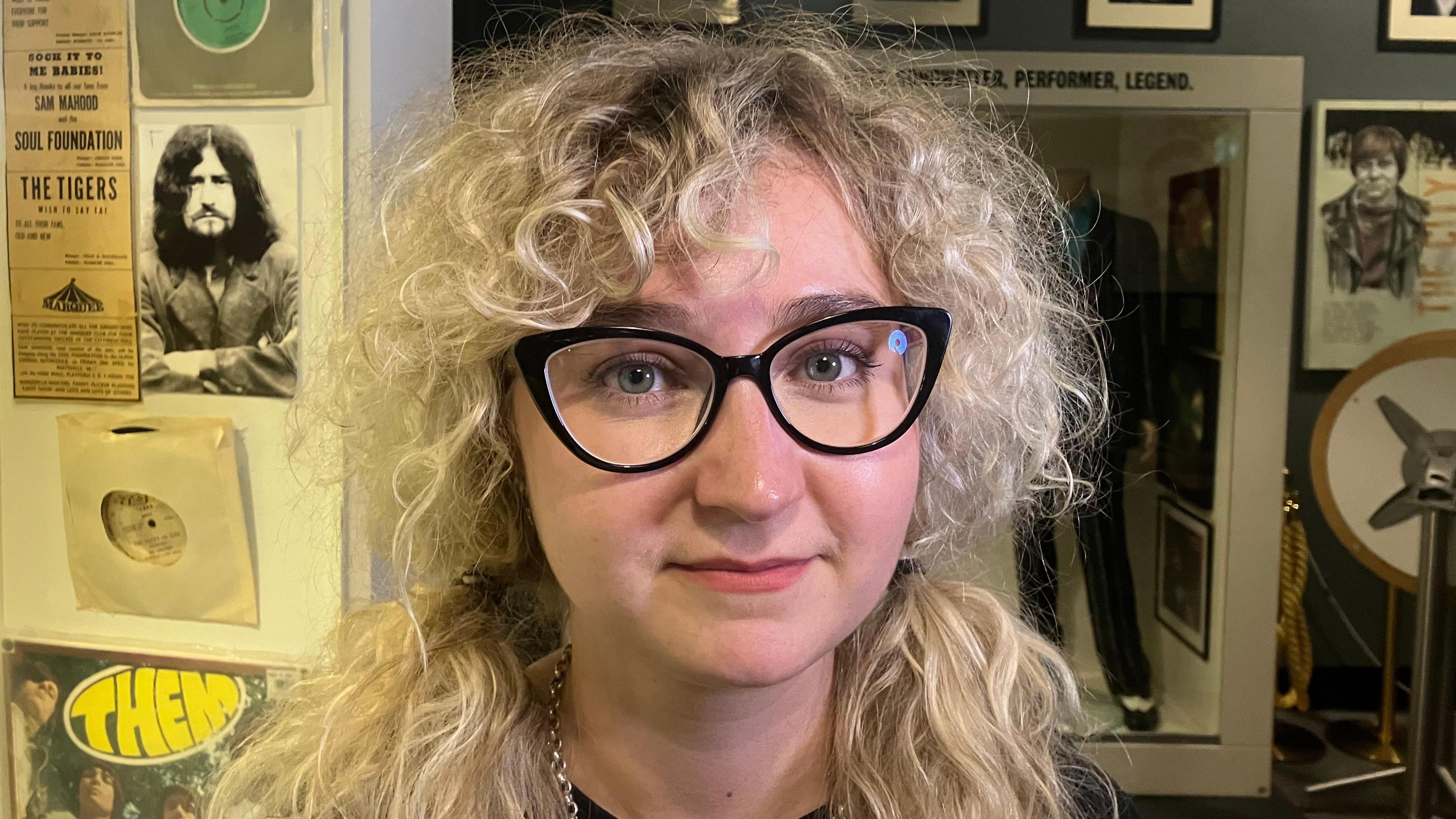 A woman with long, blonde, curly hair. She is wearing black glasses. 