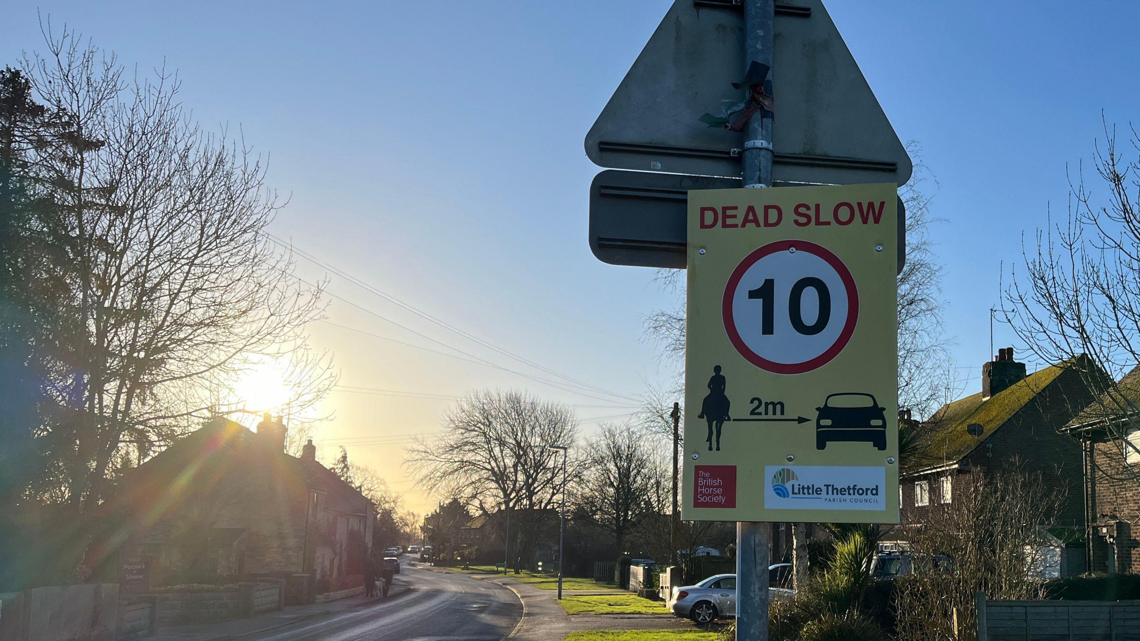 The sun rises to the left of a new yellow street sign in a residential area. The sign, situated next the the road, reads 'Dead Slow - 10'. It signals for a 2m gap to be left between horses and cars. It also feature logos from Little Thetford Parish Council and The British Horse Society. 