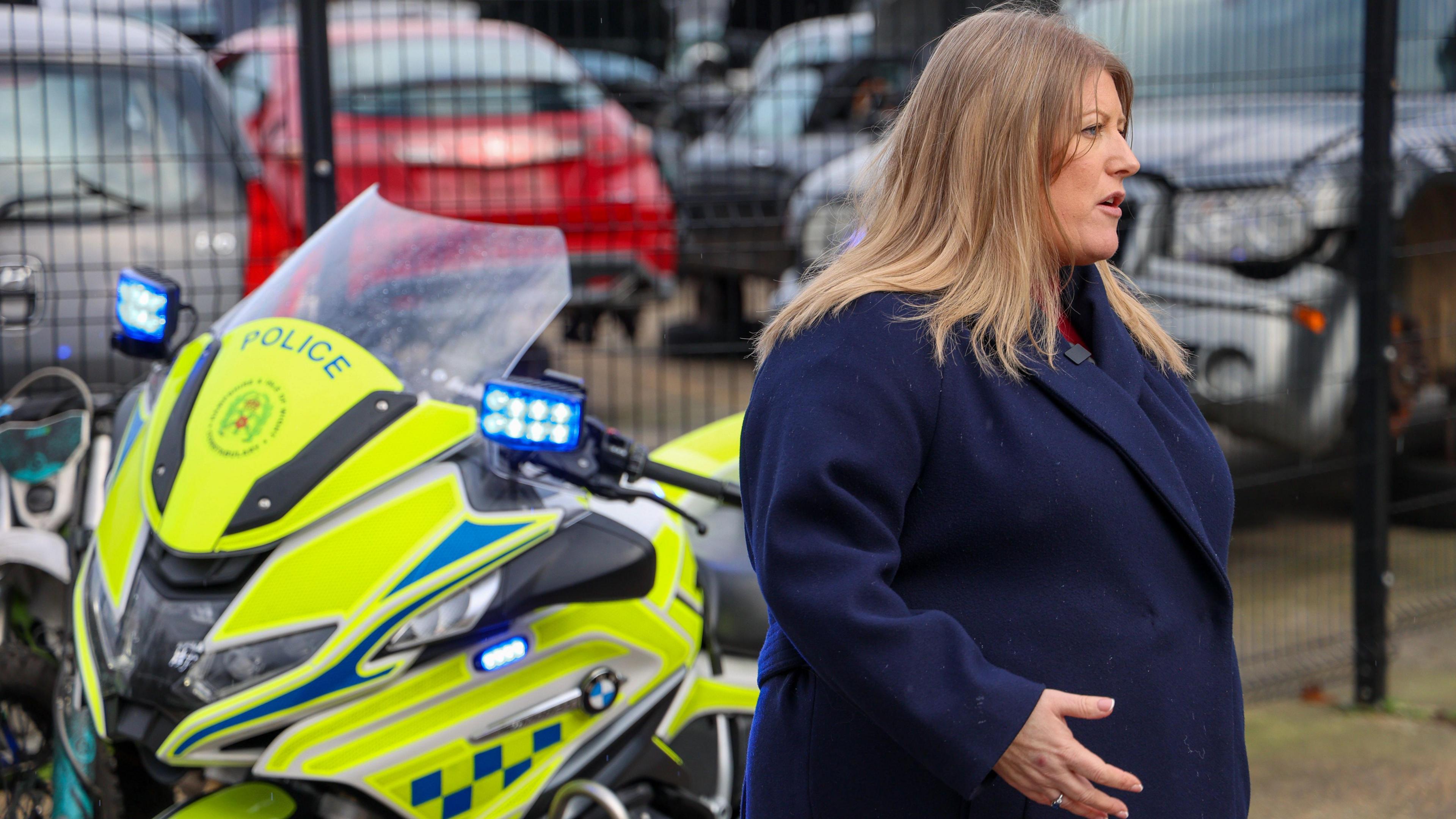 Police and Crime Commissioner Donna Jones stands in a car park, there is a black metal fence in the background with a number of cars parked up. Donna Jones stands in front of the fence, she is facing away from the camera, talking and there is a high-vis yellow police motorbike behind her. Donna Jones wears a navy blue smart woollen coat and has mid-length blonde hair.