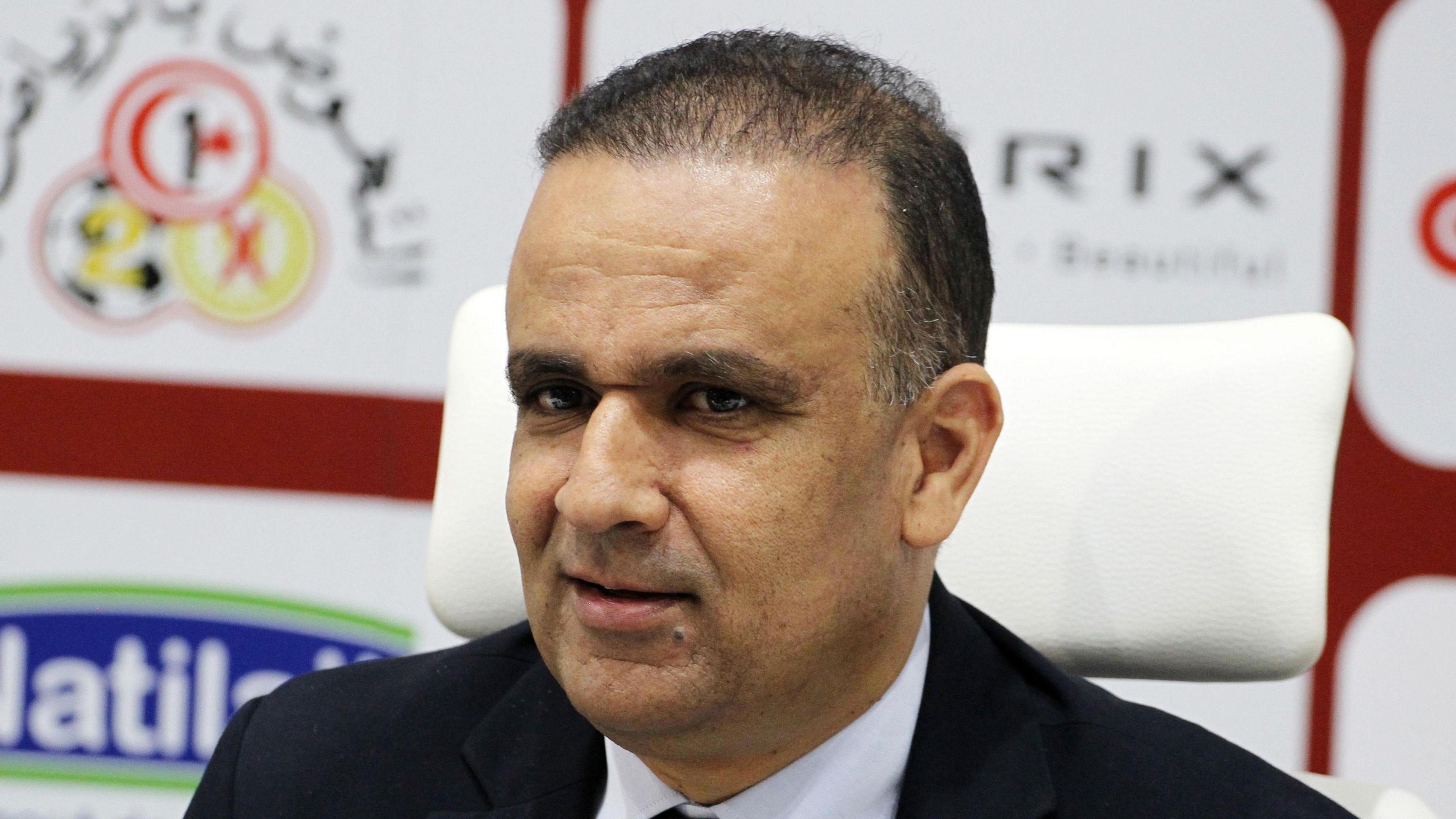 A headshot of Wadie Jary wearing a black suit jacket and white shirt, with a sponsors' advertising board behind him in the background.