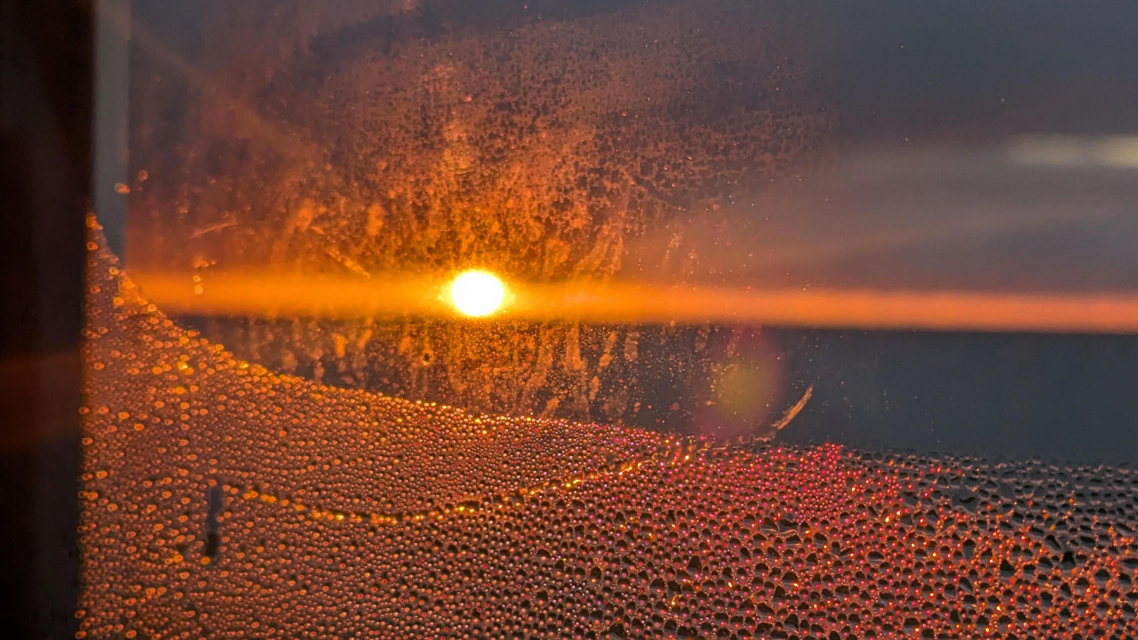 Condensation is seen covering the lower part of a window as the sun rises in the background over the sea 