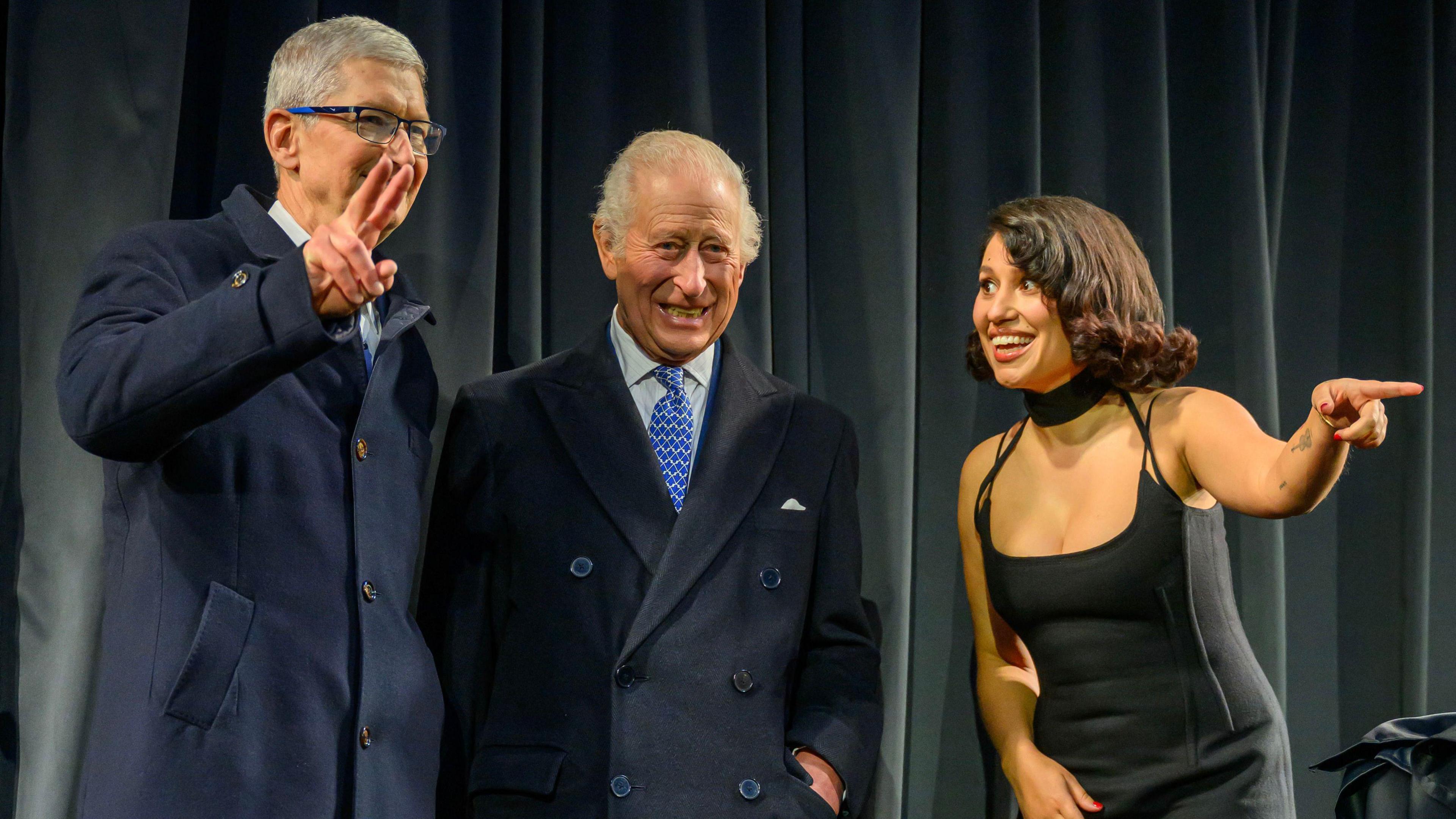 The King standing with Apple boss Tim Cook, who is making a peace sign, and Raye, who is pointing out into the audience