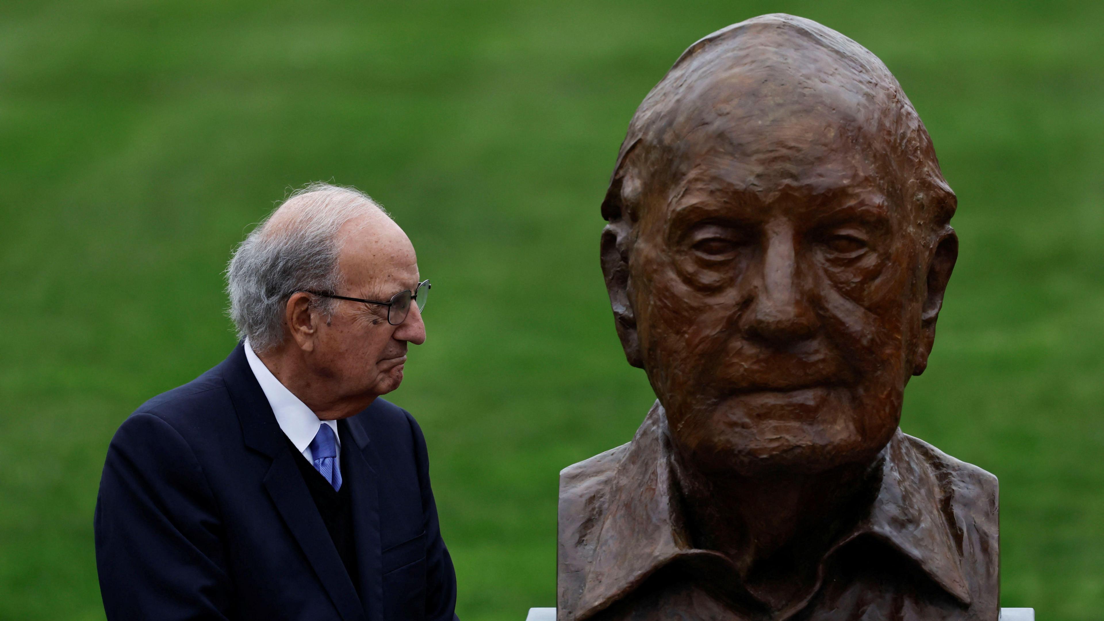 George Mitchell, with short grey hair, wearing glasses, a white shirt, purple tie and navy suit. He is looking to the right at a bust depicting himself.