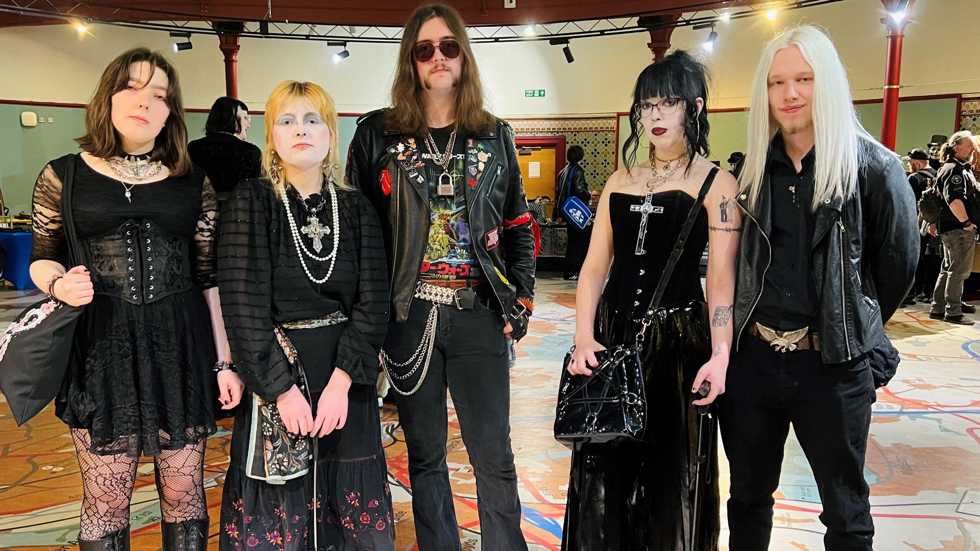 Three women and two men stood in row looking straight ahead. They are all wearing black clothing. The men have long hair while two of the women are wearing heavy make-up. They are stood in a large room with people in the background.