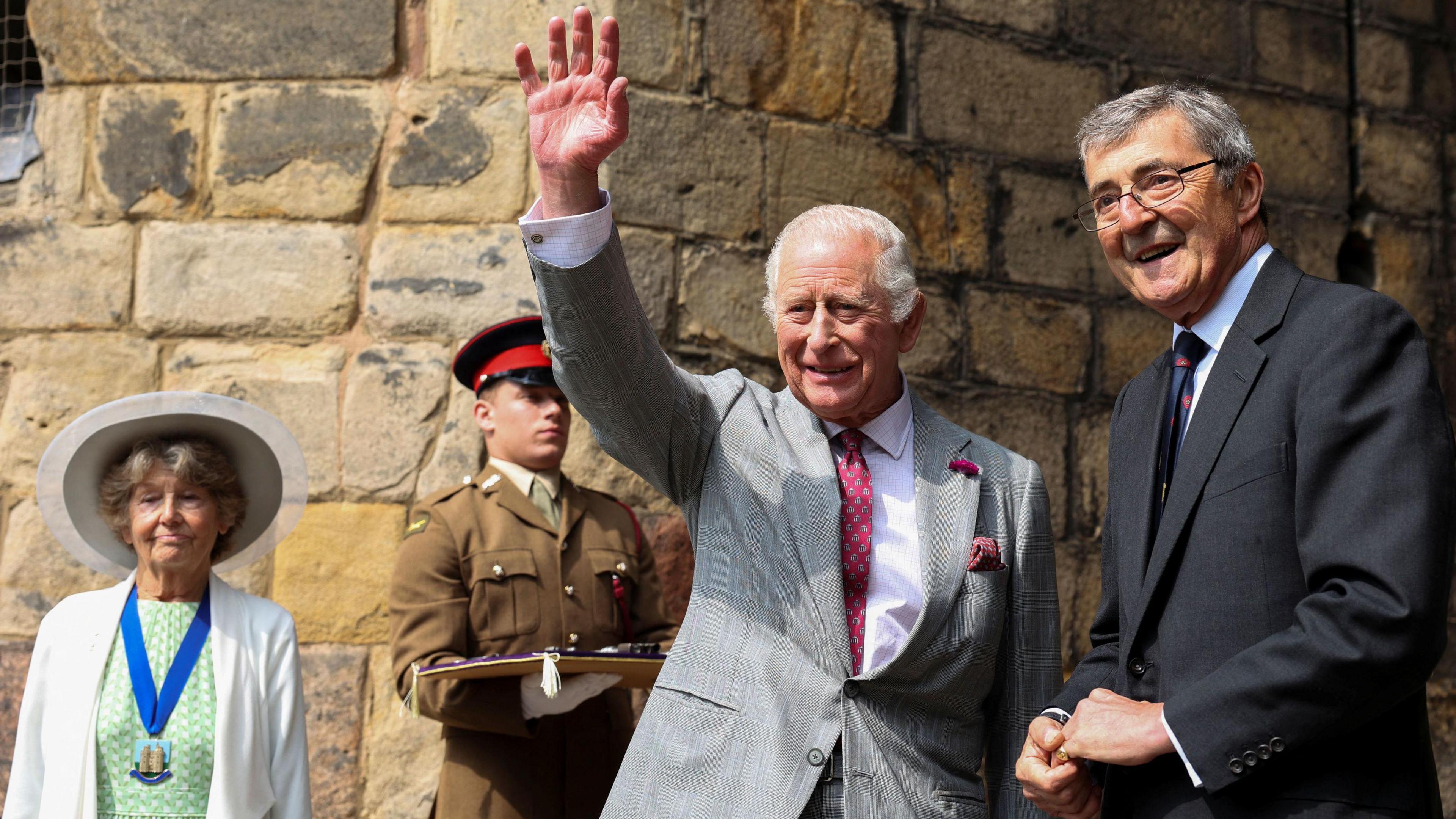 King Charles wears a light grey blazer and maroon patterned tie. He holds his arm up and waves to the crowd.