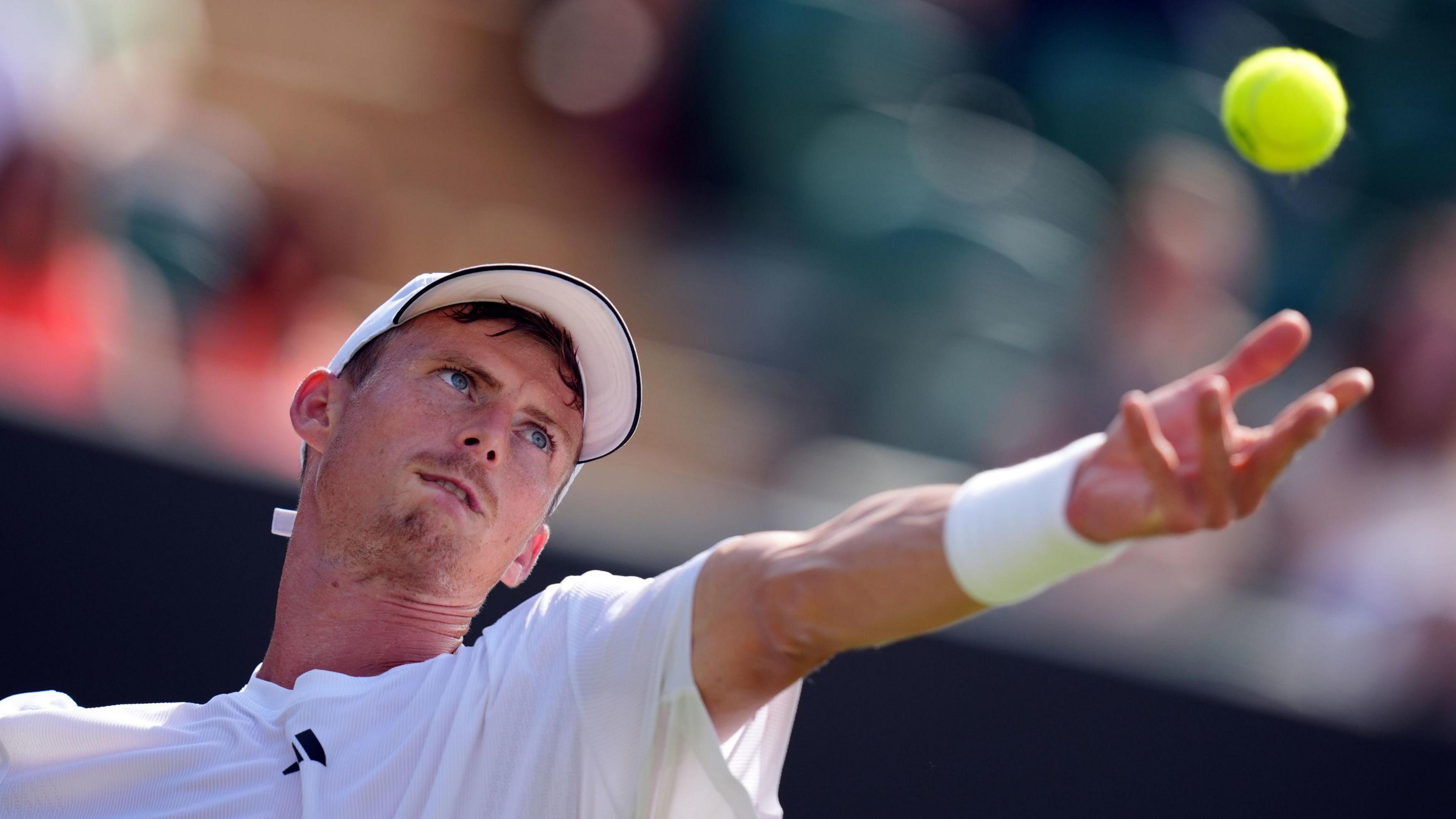 Billy Harris, who is wearing a white T-shirt, cap and wristband, throwing a ball into the air to serve. His left arm is outstretched and he has a look of concentration on his face as he focuses on the ball.
