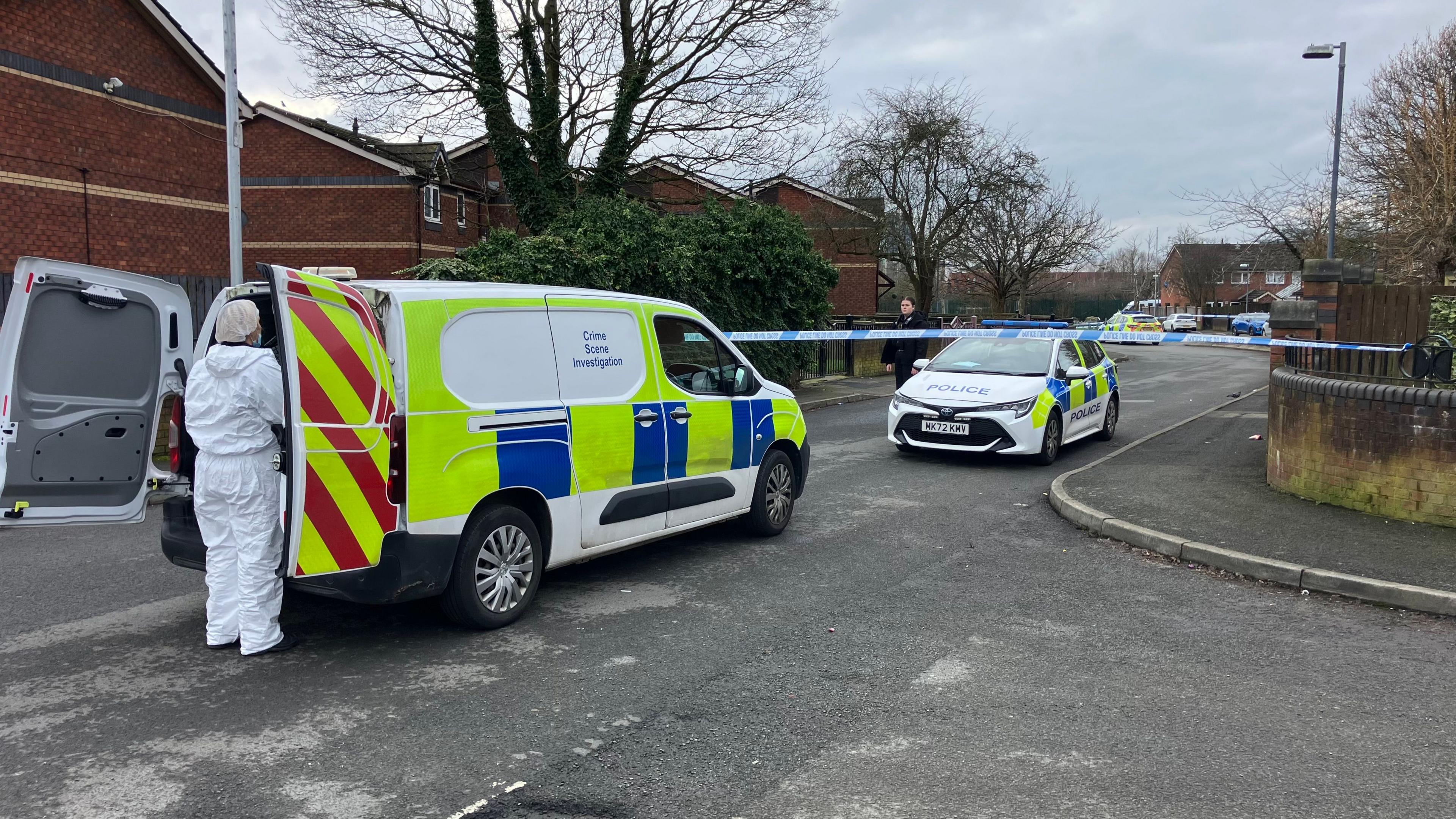 One police car and one police van can be seen on the road, where police tape has blocked off part of the residential street. A person in a full white forensics suit is in the foreground of the picture wearing a mask and head covering. 