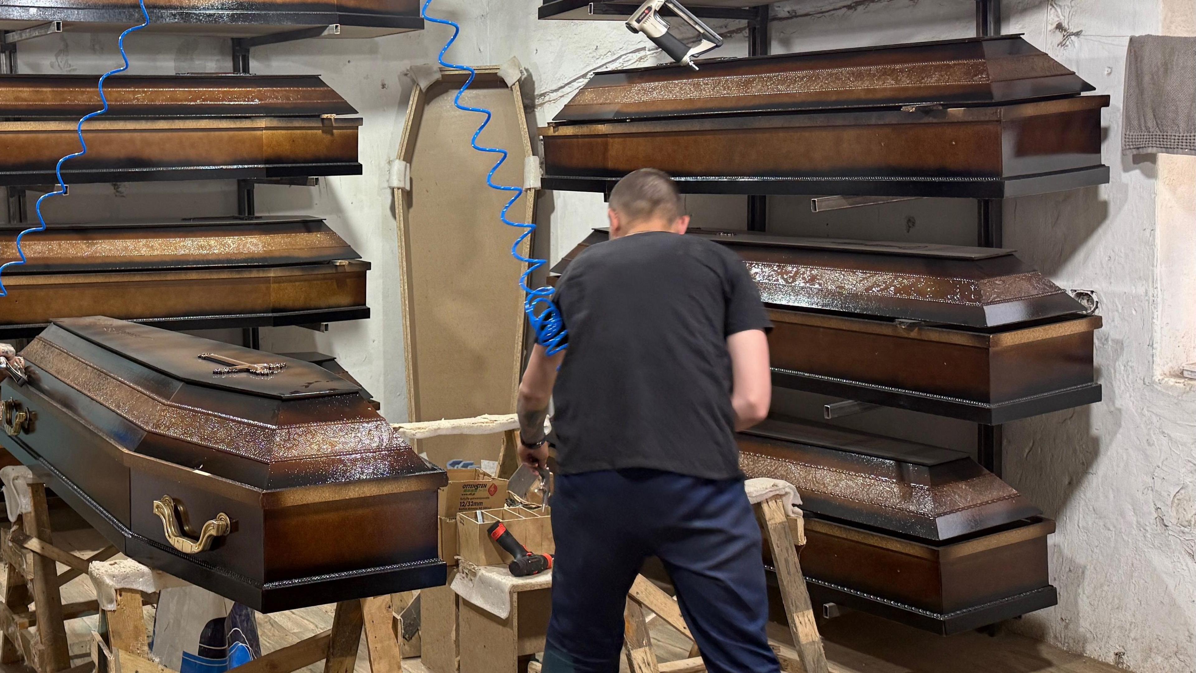 The back of a man using power tools to make coffins. There are around half a dozen identical coffins surrounding him stacked on the wall. 