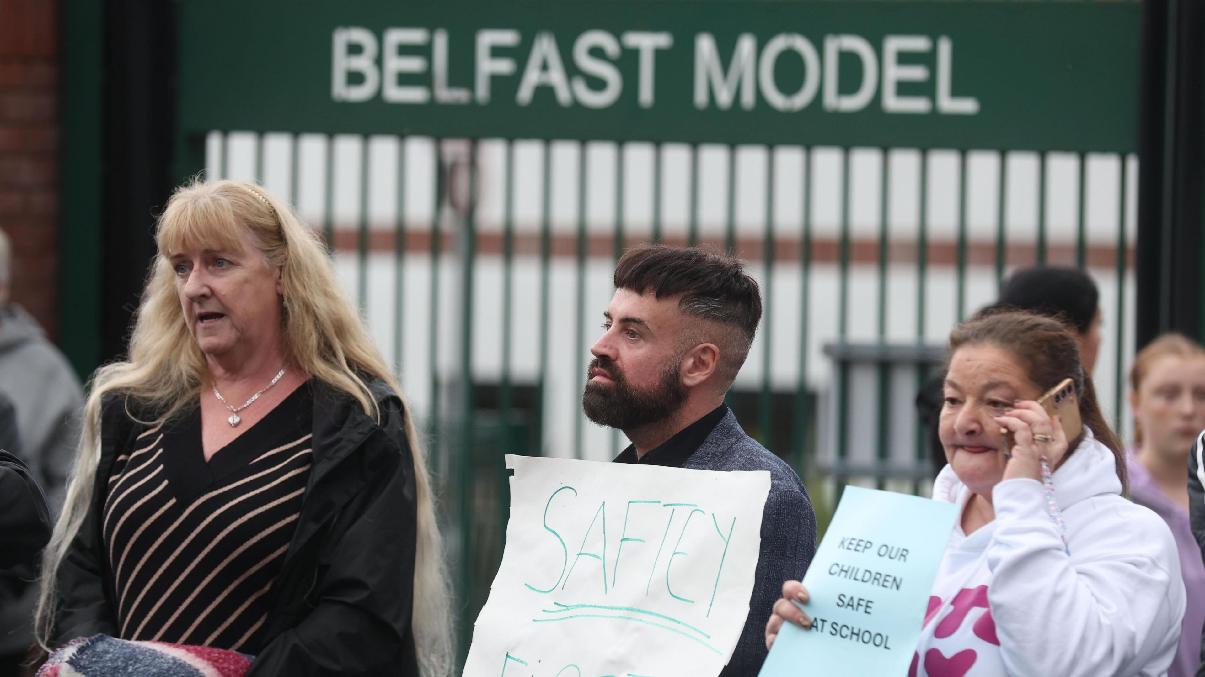 Three parents/ guardians are pictured with two holding posters. The first lady with long blonde hair appears to be speaking to someone. Meanwhile the man and other woman are holding signs.