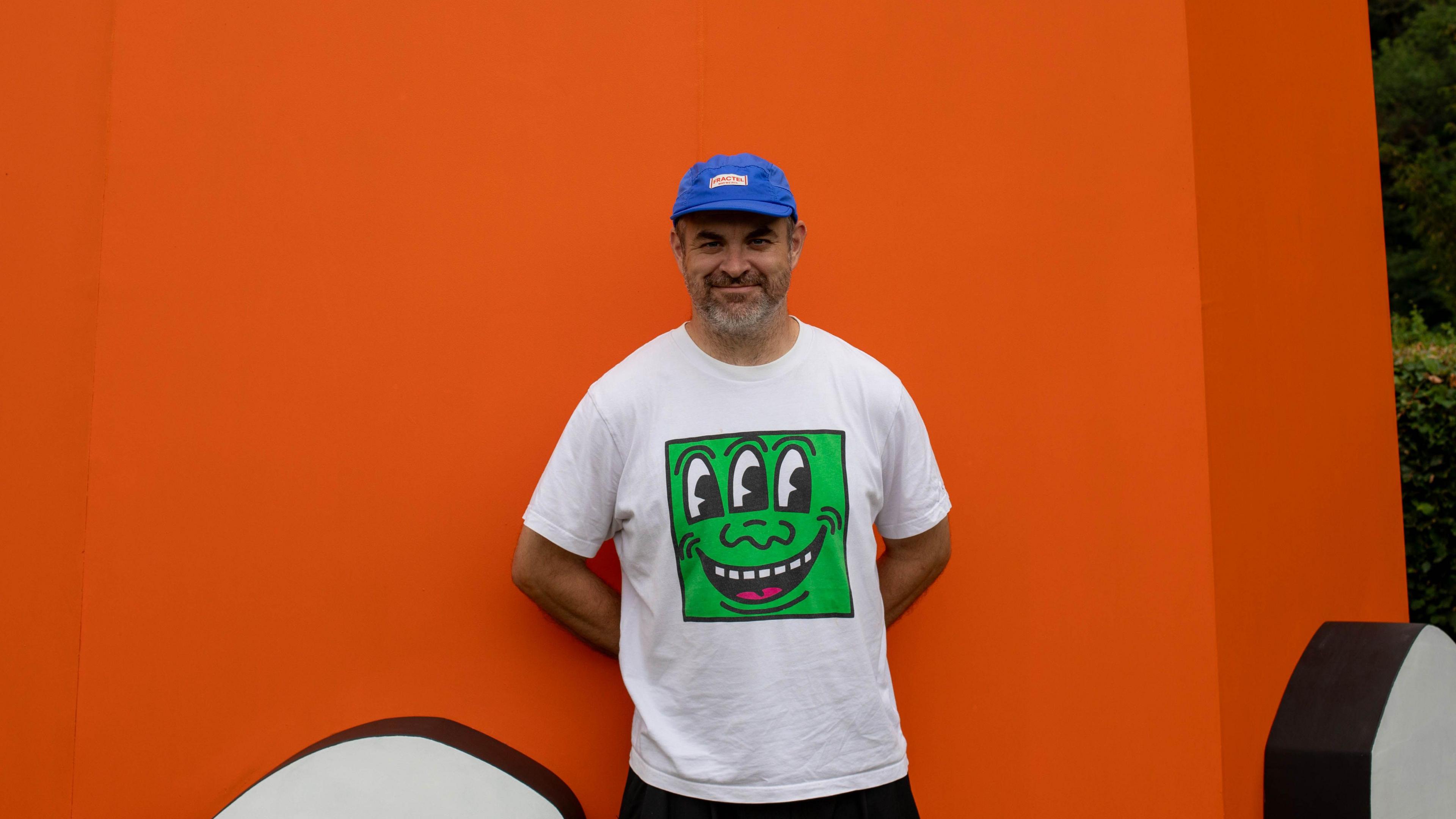 A man wearing a white T-shirt and a blue baseball cap is standing in front of a bright orange sculpture. He is smiling and looking at the camera.