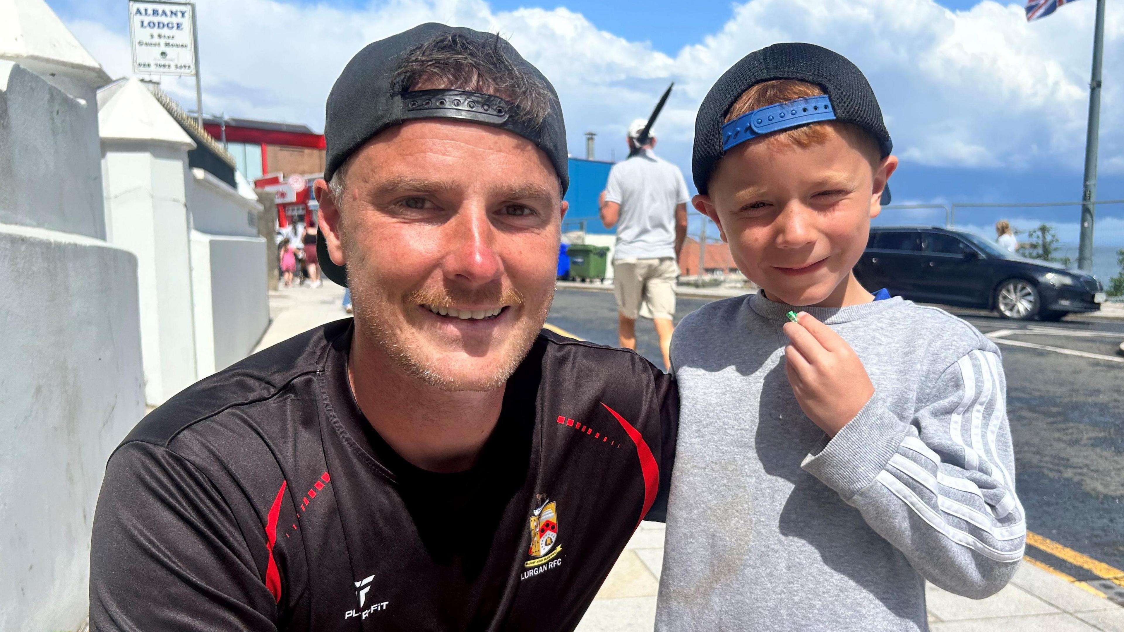Simon Patterson and his son Sam are both smiling as Simon, on the left of the image, kneels down and son Sam stands. Simon is wearing a black baseball cap and black football team shirt. Sam has one hand up to his mouth and is wearing a grey sweatshirt and baseball cap. Both Simon and sam have their caps turned backwards.